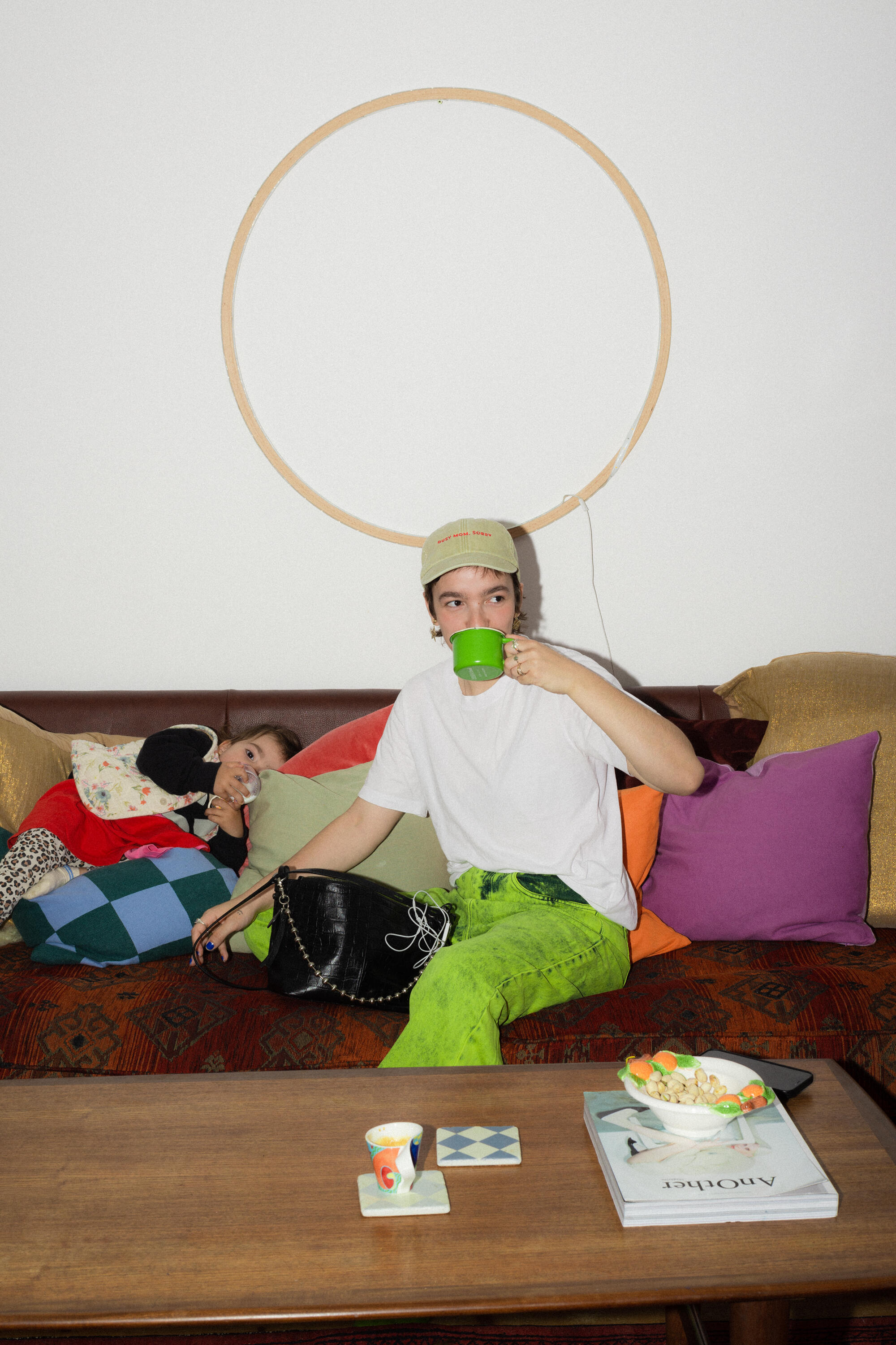 A person in a green outfit sits on a couch holding a circular INA KENT frame, with another person reclining beside them. A coffee table with items and colorful pillows are visible.