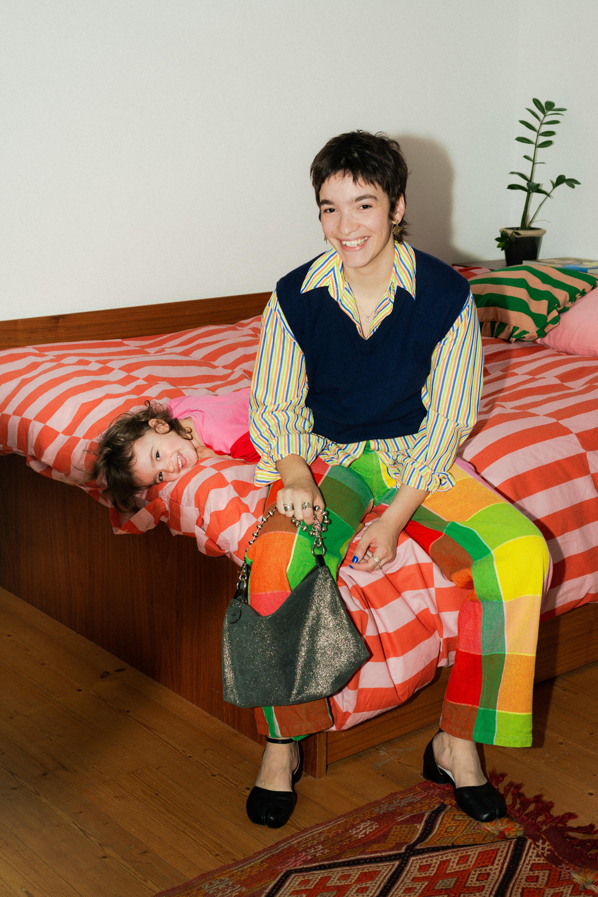 A smiling adult sitting on a bed with a playful child beside them, both dressed in colorful clothing, in a room with wooden flooring and a plant, holding a bag from INA KENT.