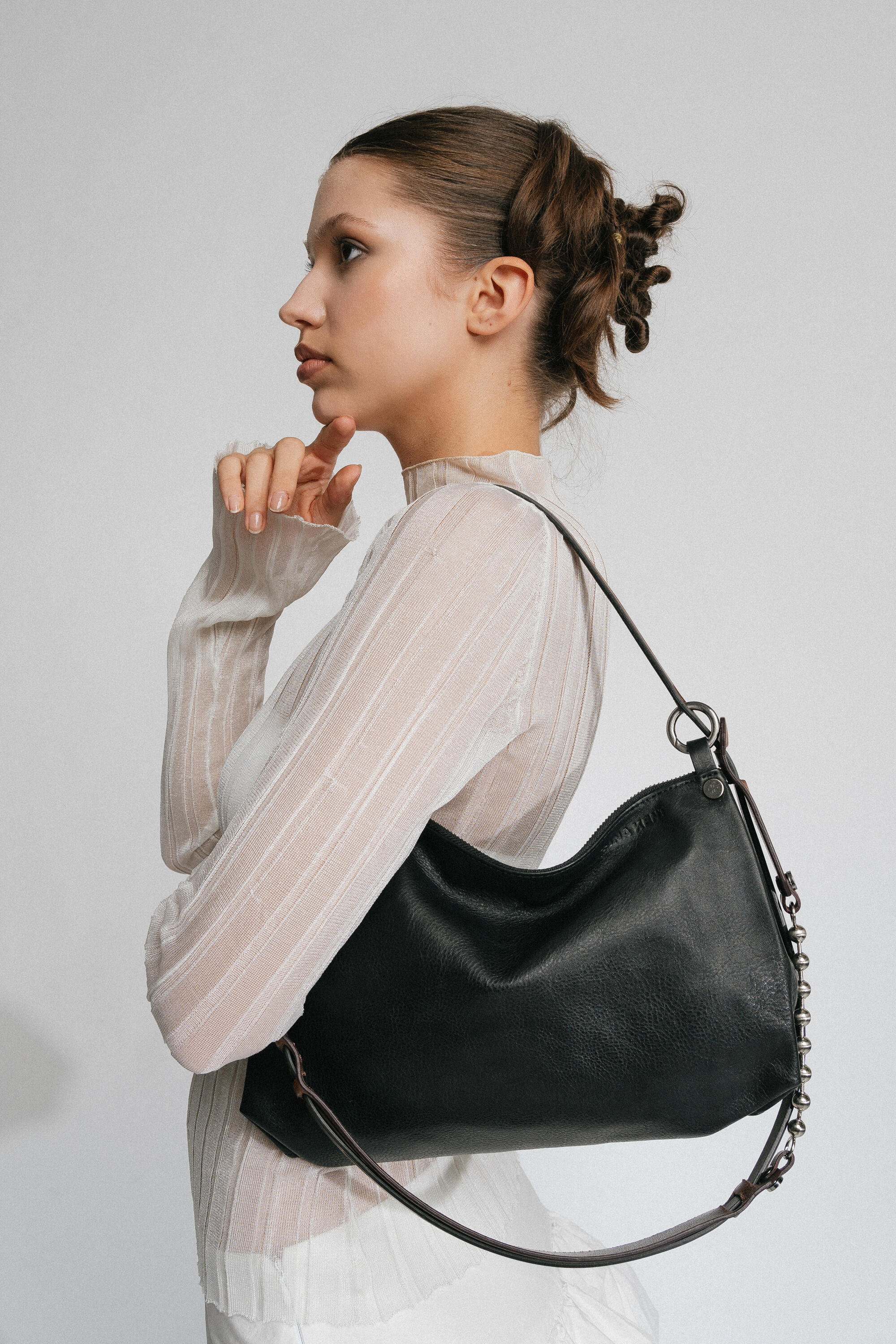 A woman in a white long-sleeve top poses in profile with her hand under her chin, wearing an INA KENT black leather shoulder bag with a chain strap.