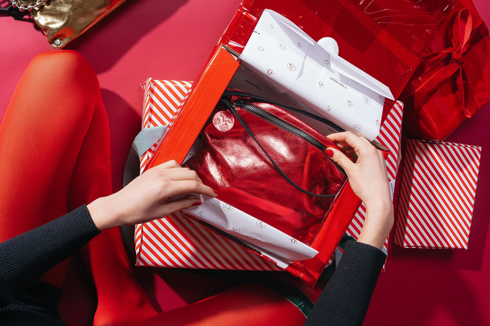 Person in roter Strumpfhose und schwarzem Oberteil öffnet ein rot-weiß-gestreiftes Geschenkpapier, in dem sich die Tasche MOONLIT ed.1 in der Farbe Eco Antique Metallic Chili Red befindet. Die glänzende, metallisch rote Tasche liegt in einer roten Geschenkbox, umgeben von weiteren rot-weiß-gestreiften und roten Geschenkpaketen.