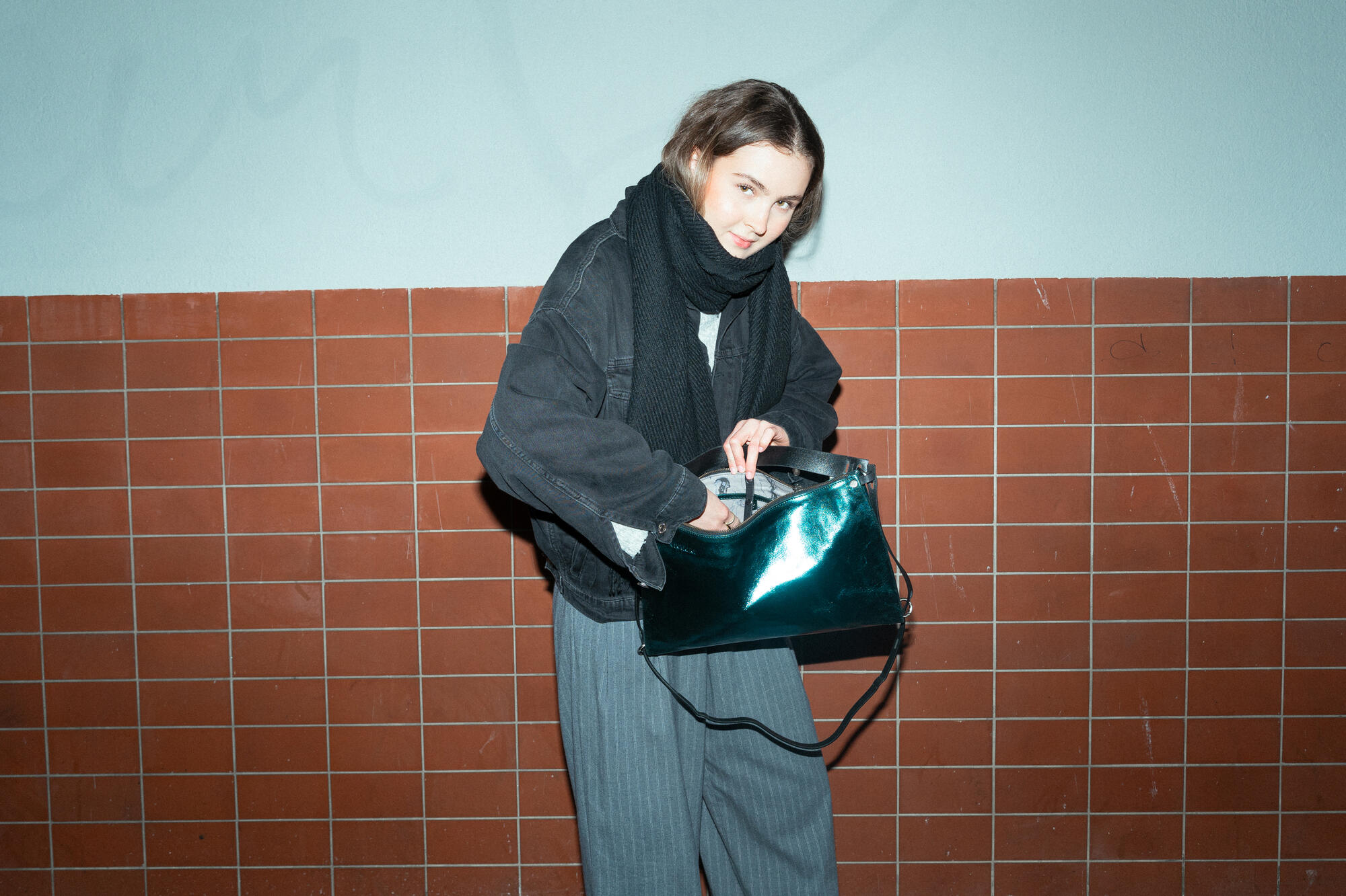 A female-presenting person stands in front of an orange-brown tiled wall and looks down slightly as she reaches into her shiny, dark green bag (“DINKUM ed.2” model in crackled dark teal leather). She is wearing a black jacket, a thick scarf and wide gray pants. 