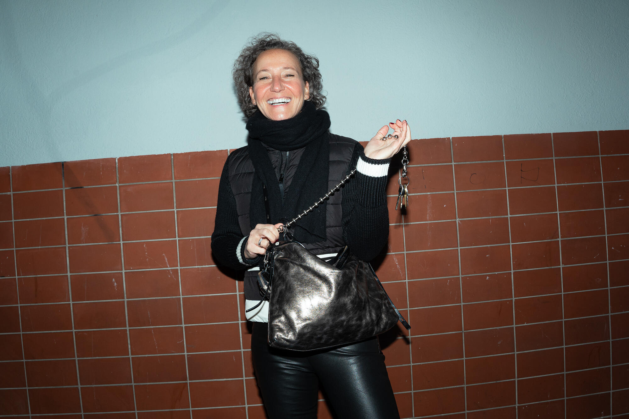 A female-presenting person is wearing a black jacket, a black scarf and black leather pants. She is holding the “ROVE ed.1” bag in metallic dark anthra. She is also holding a bunch of keys in her hand and smiling broadly.