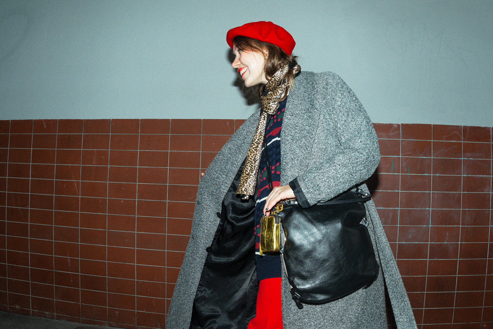 A female-presenting person is wearing a gray coat, a red beret and a leopard-print scarf. She is holding the black bag by INA KENT “AD LIB ed.1”, to which a shiny gold-colored pendant is attached. 