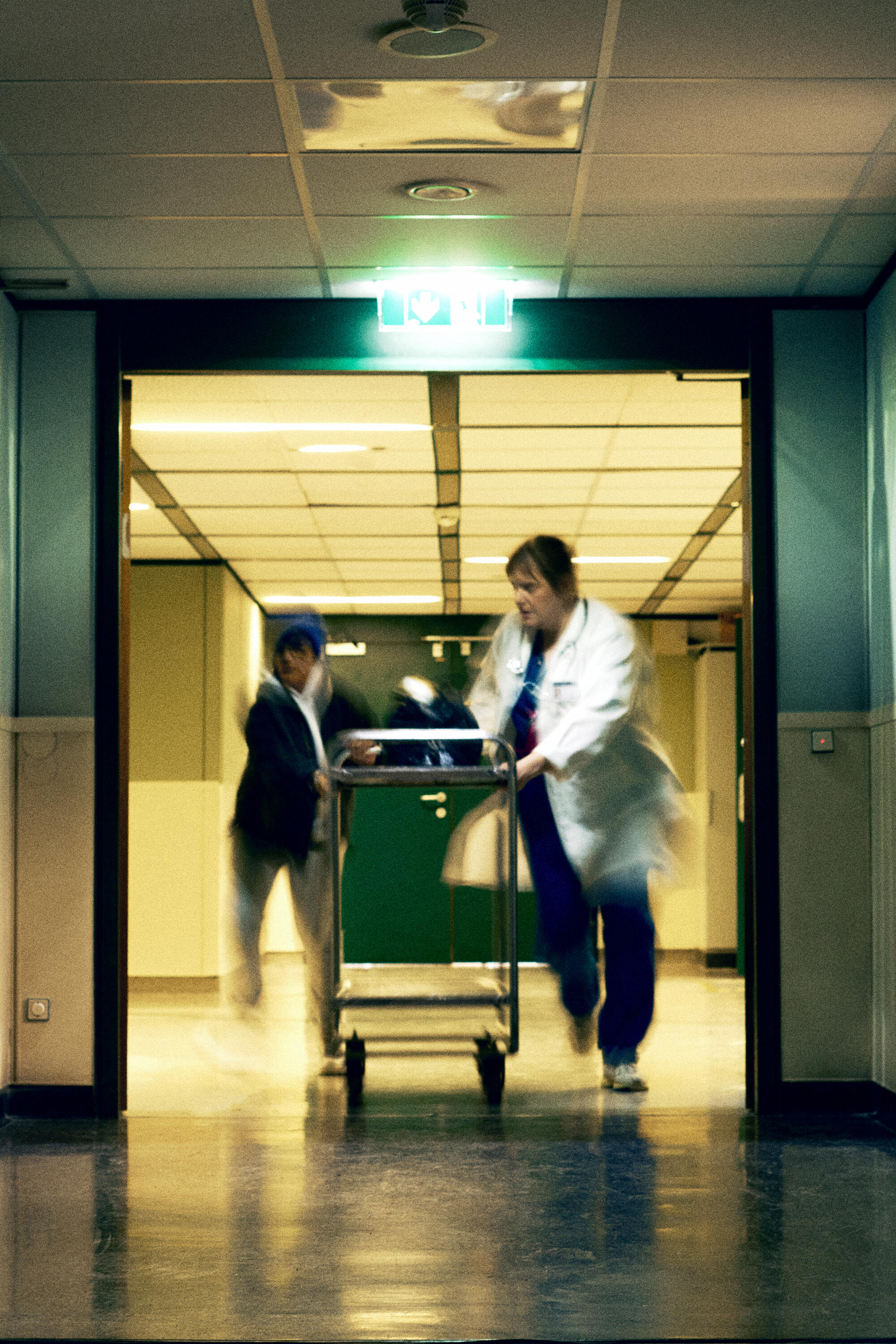 Two medical professionals rush a patient on a gurney down a hospital corridor under an illuminated emergency exit sign, while one carries an INA KENT bag over their shoulder.