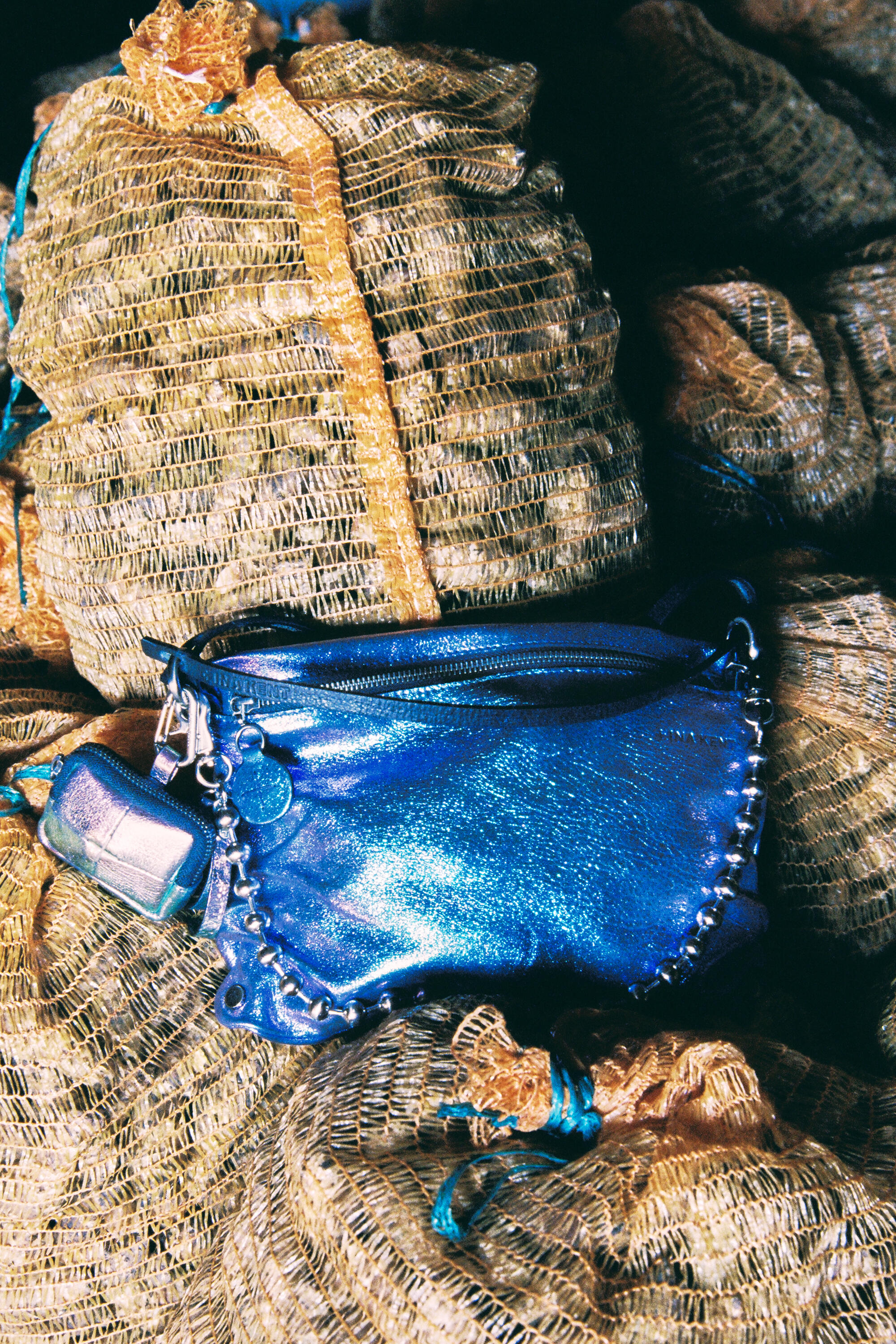 Eine glänzend blaue INA KENT-Handtasche mit Kettendetail liegt auf gestapelten Netztaschen voller Produkte.