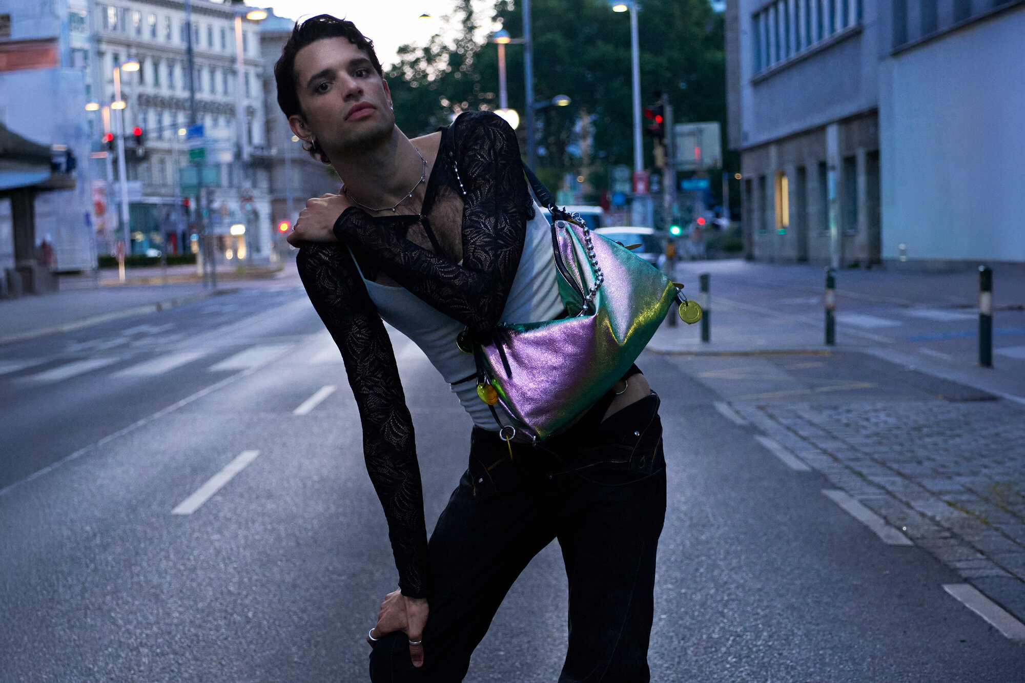 A person poses in the middle of an empty city street at dusk, wearing a sheer black top, dark pants, and carrying a shiny iridescent INA KENT bag.