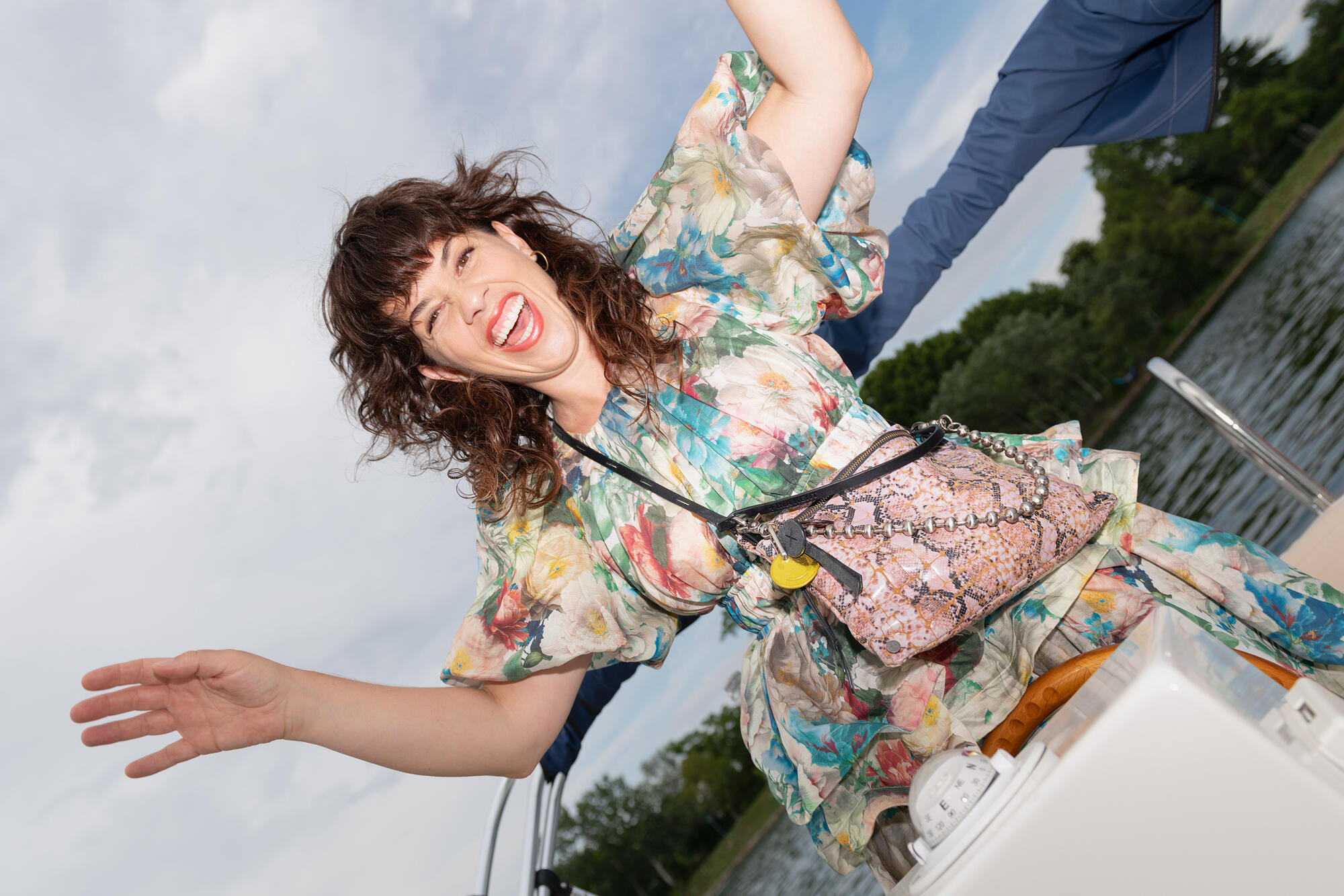 Eine Frau in einem geblümten Kleid, die eine INA KENT-Tasche trägt, lächelt und posiert spielerisch auf einem Boot, mit Wasser und Bäumen im Hintergrund unter einem bewölkten Himmel.