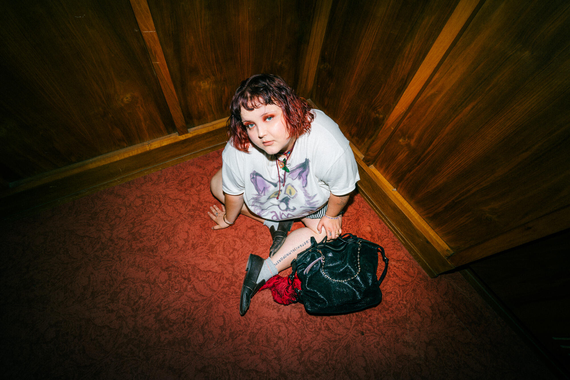 Person with short red hair sits on a carpeted floor in the corner of a wood-paneled room, wearing a cat graphic t-shirt and boots, with an INA KENT black bag beside them.
