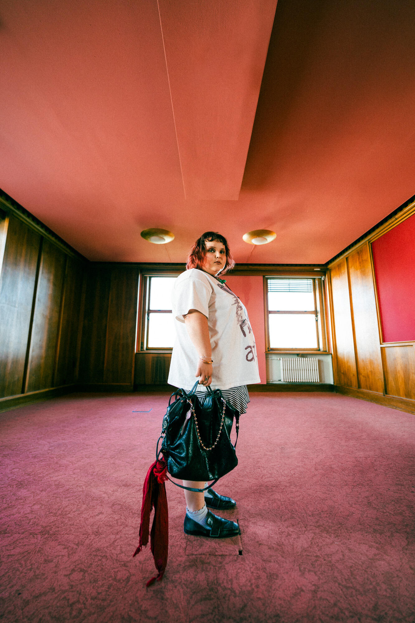 A person with short hair stands in the center of an empty room with wooden walls and a red ceiling, holding a black INA KENT bag and a red scarf.