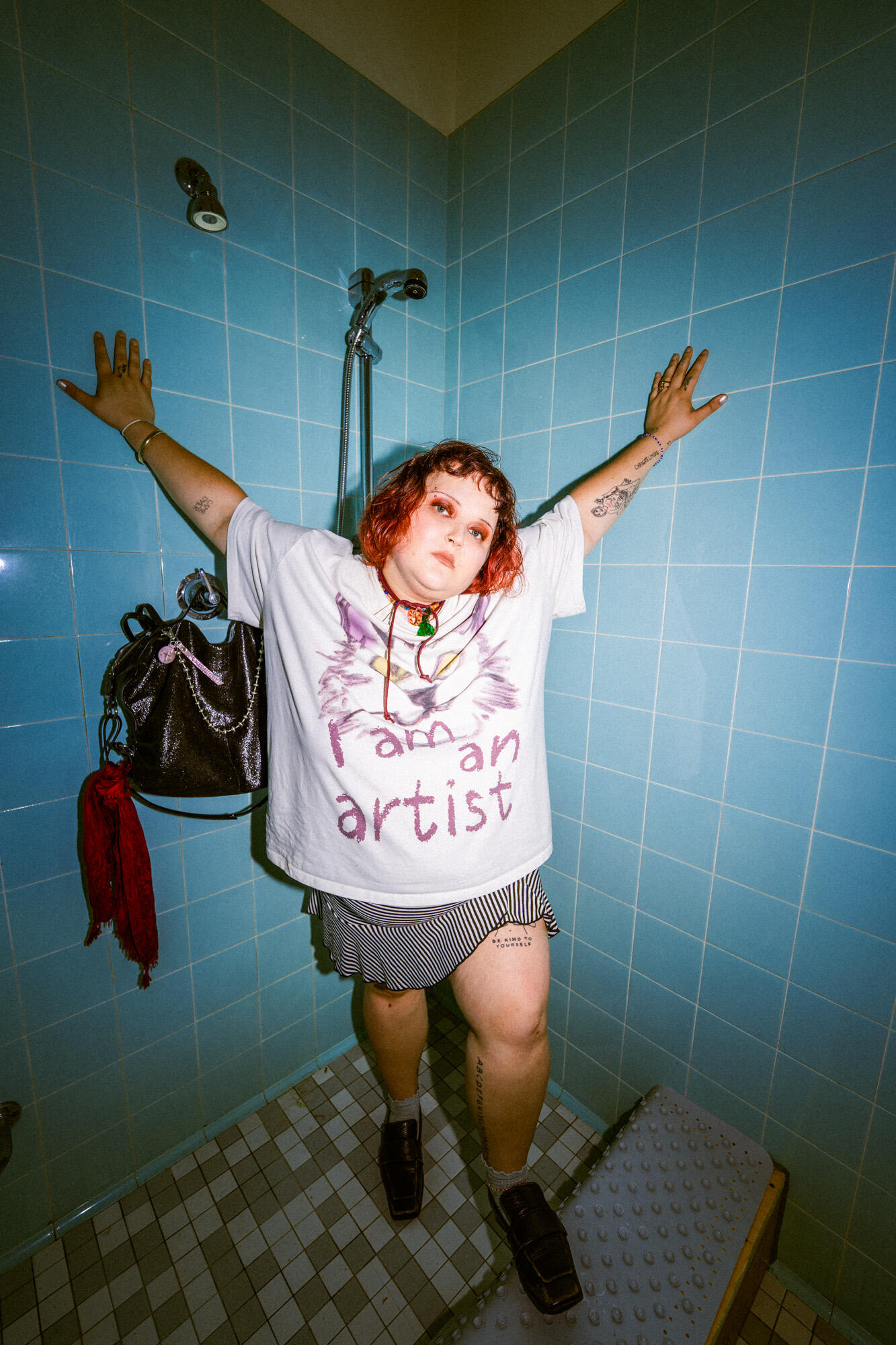 Person with short red hair and tattoos stands in a tiled shower, arms outstretched, wearing a "faman artist" t-shirt, striped shorts, and black shoes. An INA KENT black bag hangs nearby.