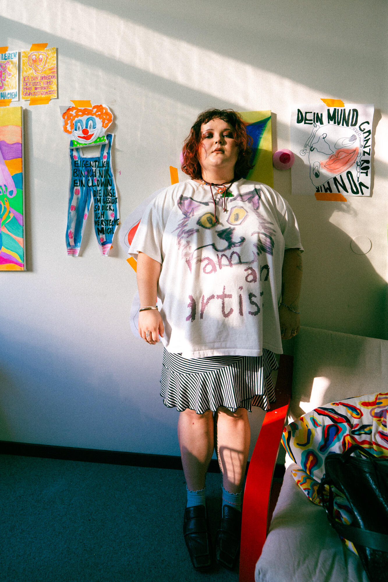 A person with short, curly hair stands in a colorful room wearing a shirt that reads "I am an artist." Behind them, artwork, posters, and an INA KENT bag add creativity to the vibrant decor.