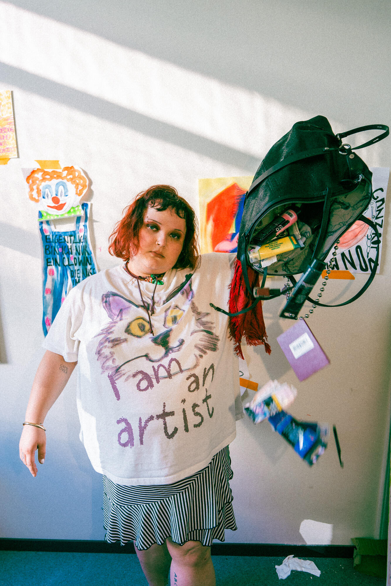 A person with red hair wearing an "I am an artist" shirt holds up an INA KENT backpack, spilling its contents. Artworks and posters are visible on the wall behind them.