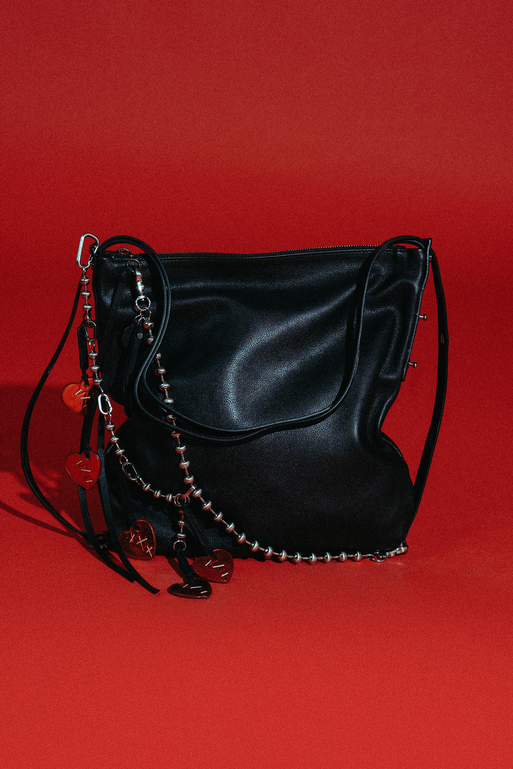 Black leather INA KENT handbag with a zipper, silver chain trim, and decorative metallic charms, displayed against a solid red background.