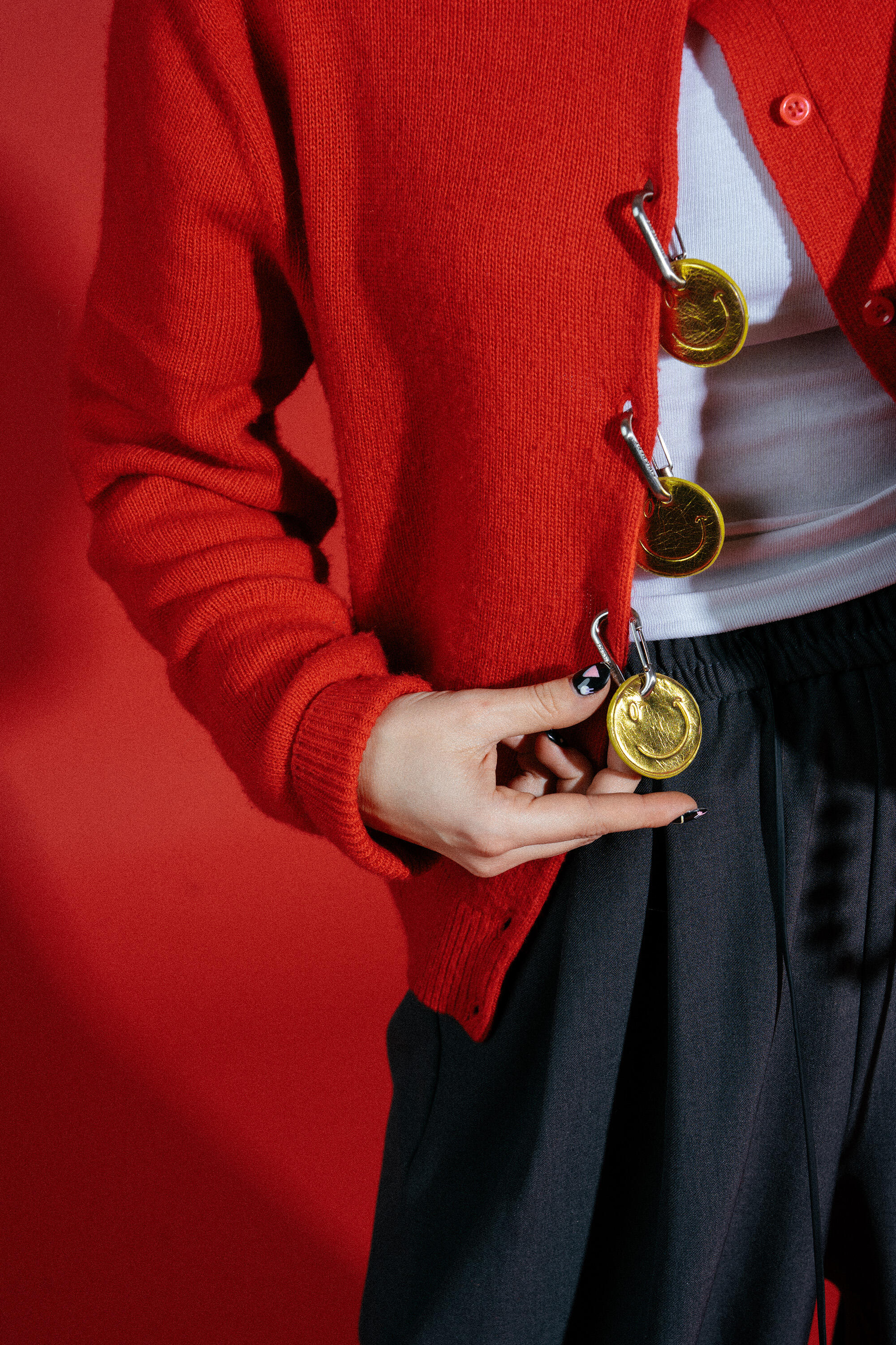 A person wearing a red cardigan fastened with large safety pins and gold medals, over a white shirt and dark trousers, stands against a red background, accessorized with an INA KENT bag.