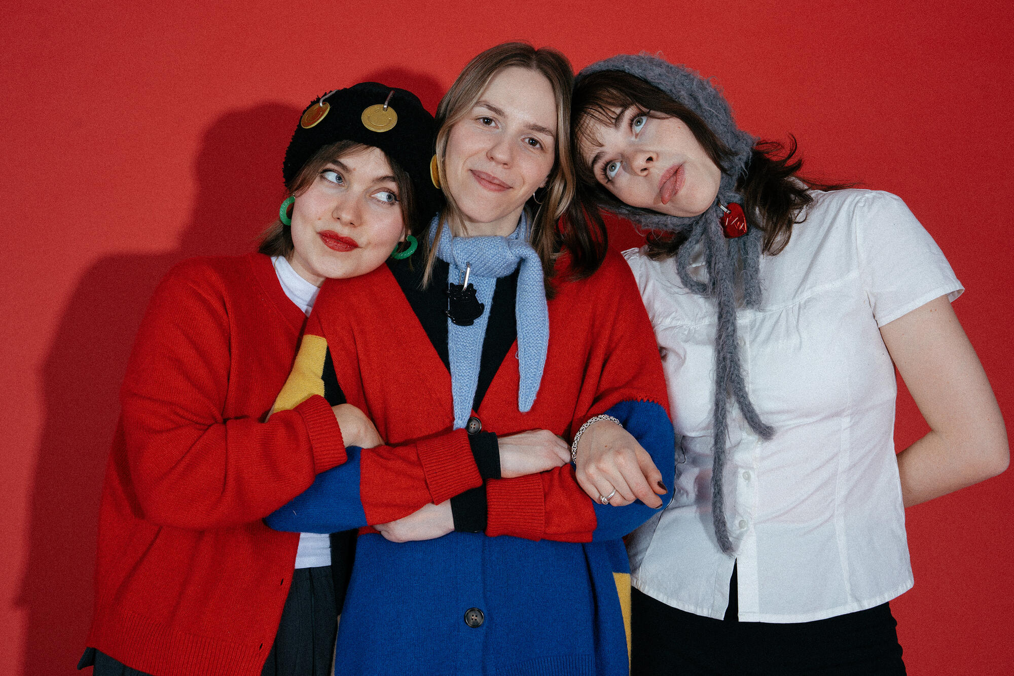 Three people stand close together against a red background, wearing colorful sweaters, hats, and neutral expressions—each carrying a stylish INA KENT bag.