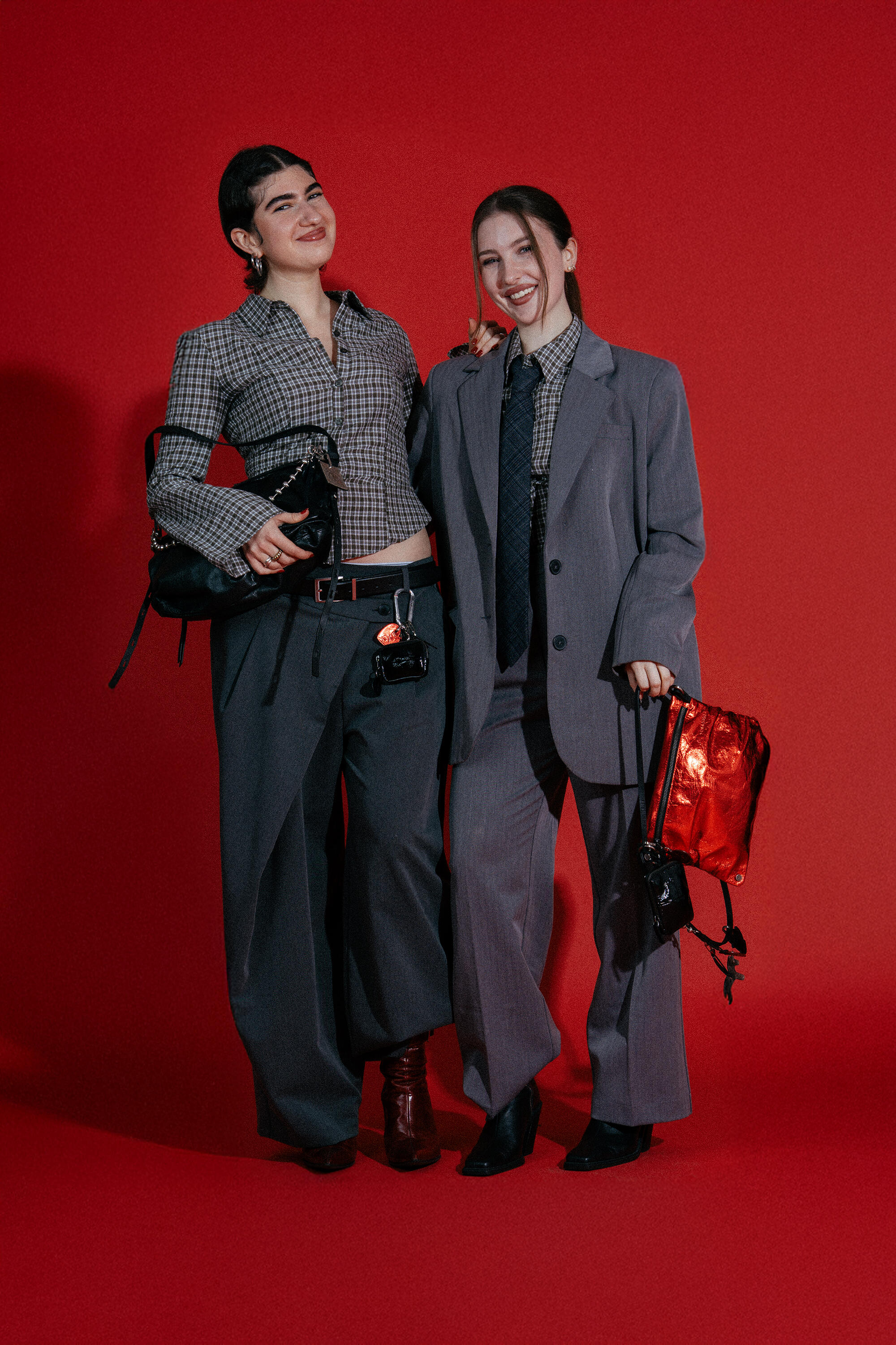 Two women dressed in oversized suits stand against a red background, smiling and holding stylish INA KENT handbags.