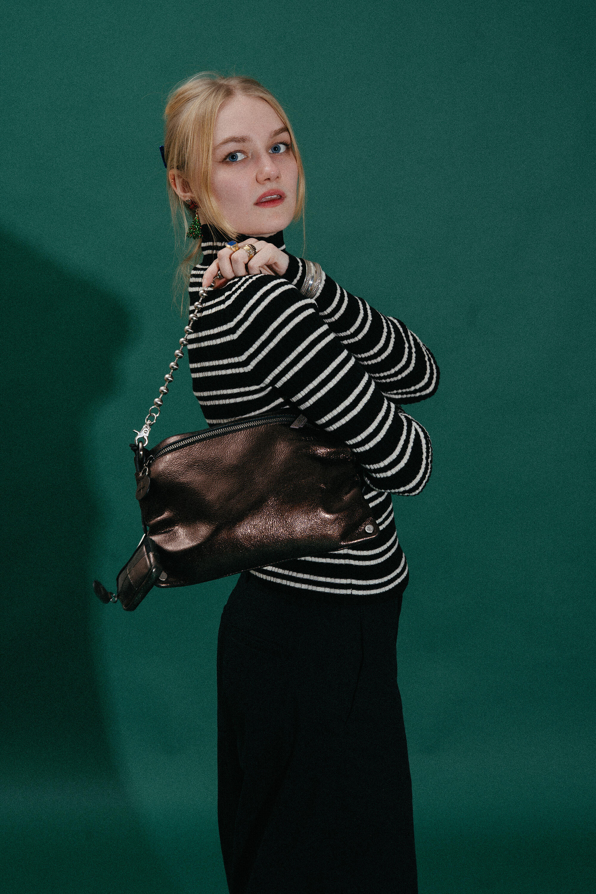 A person stands against a green background, wearing a black and white striped long-sleeve shirt, black pants, and holding an INA KENT metallic shoulder bag.