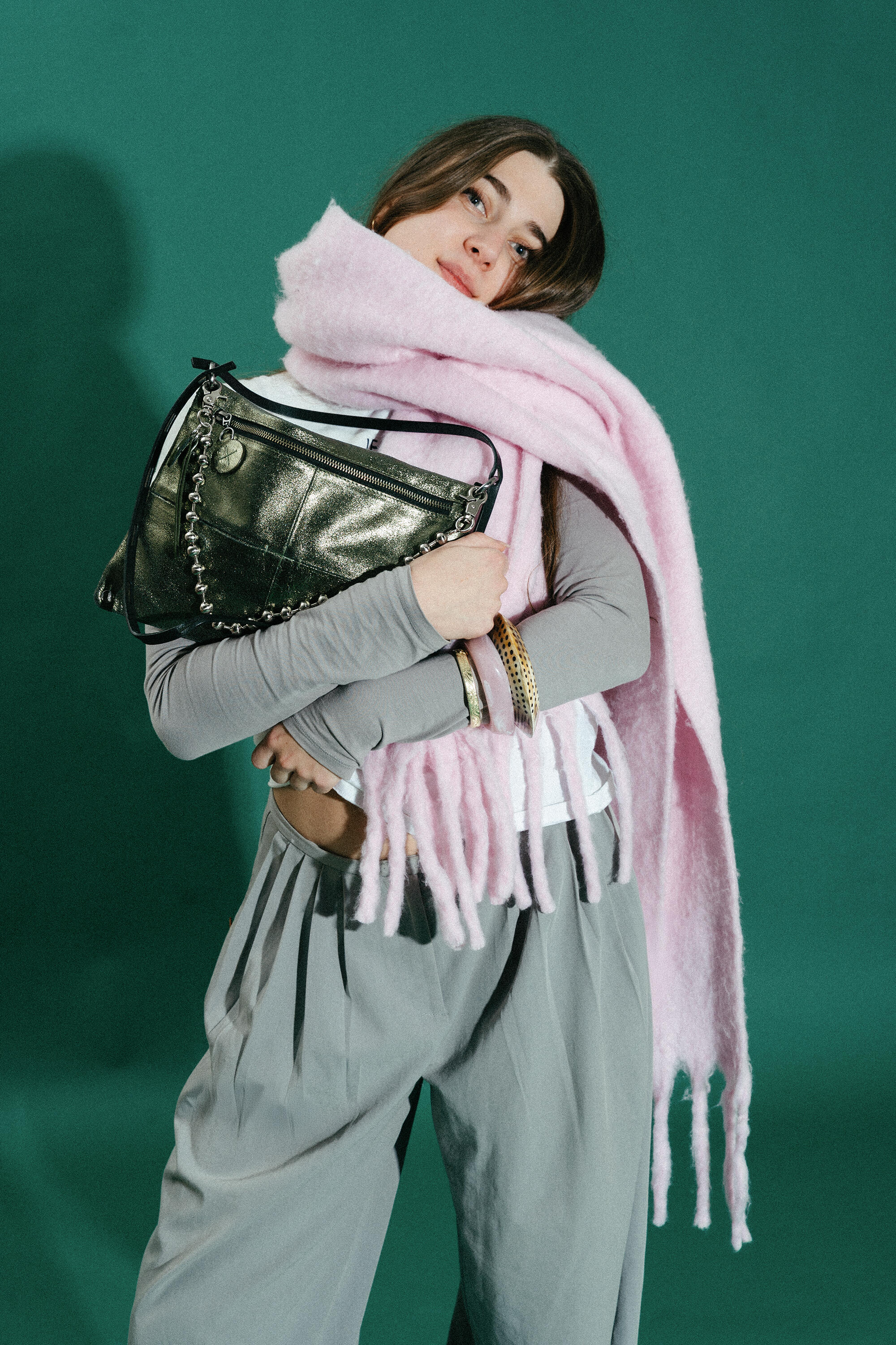 Woman standing against a green background, wearing gray pants, a gray long-sleeve top, and a large pink scarf, holding an INA KENT metallic handbag and accessorized with bracelets.