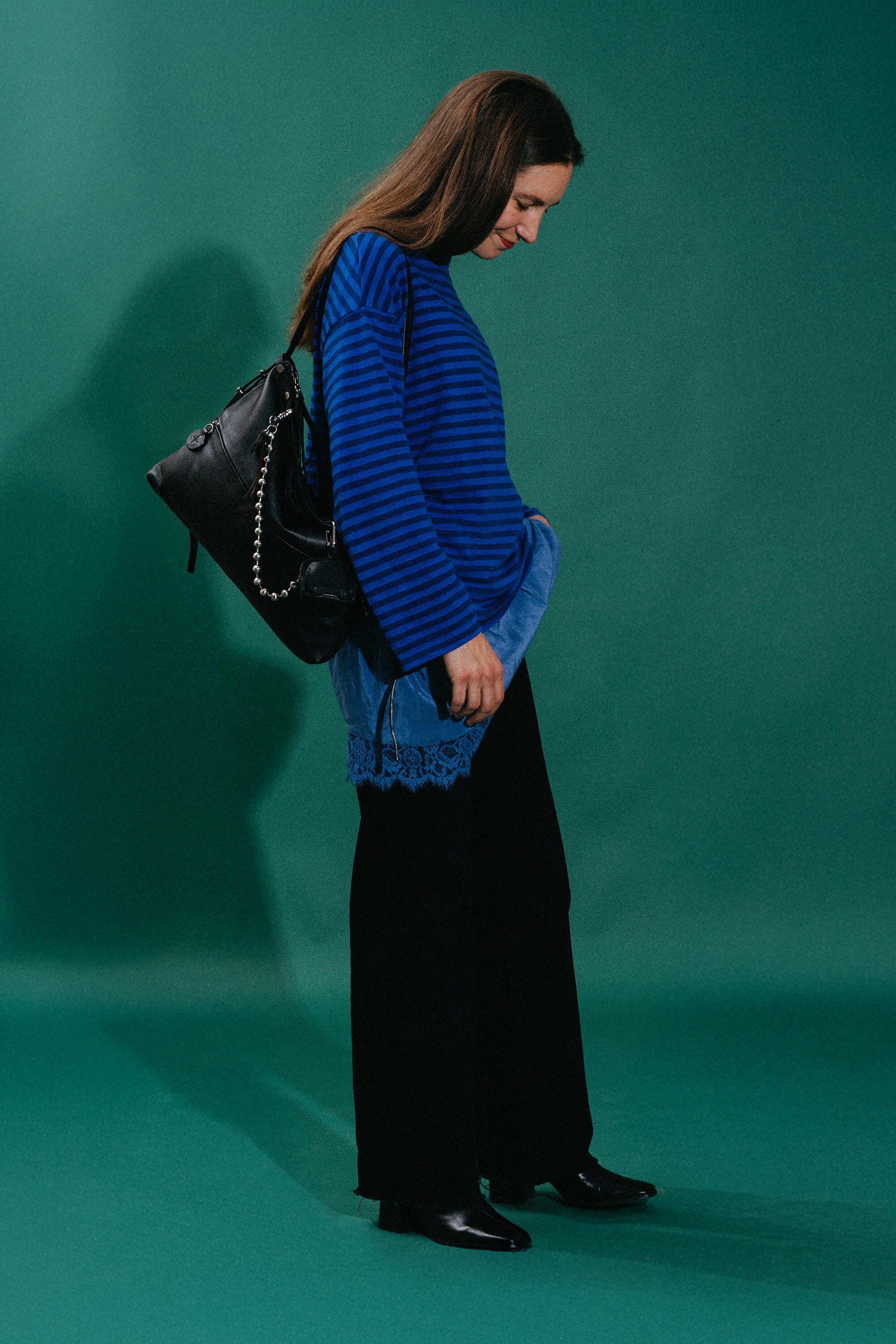 A woman stands in profile against a green backdrop, wearing a blue striped sweater, black pants, black shoes, and carrying an INA KENT black handbag over her shoulder.
