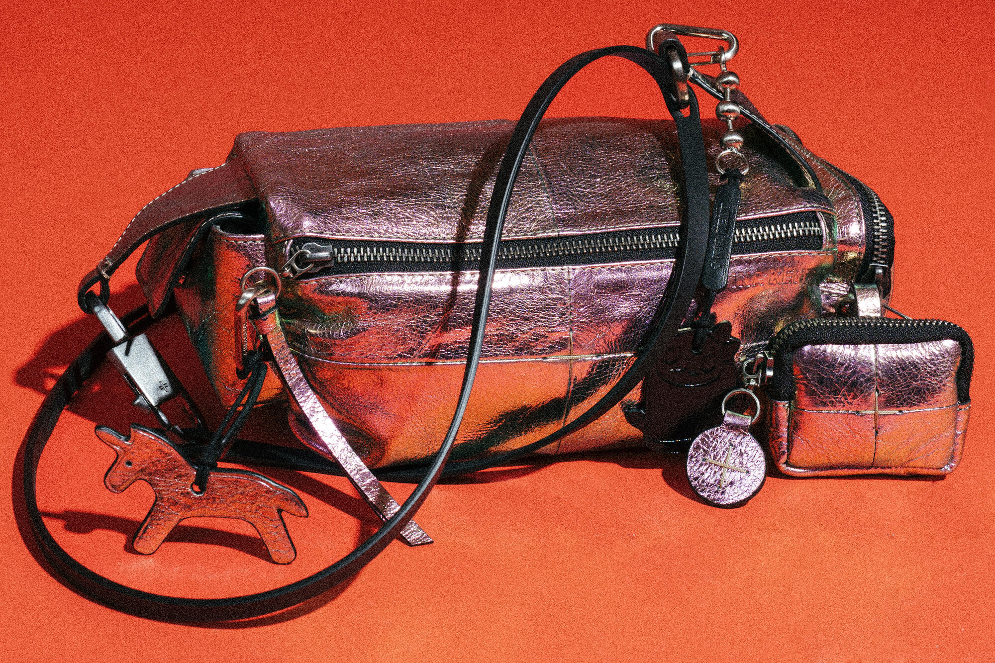 A metallic pink INA KENT handbag with a matching coin pouch, black strap, silver hardware, and a dog-shaped keychain, set against a red background.