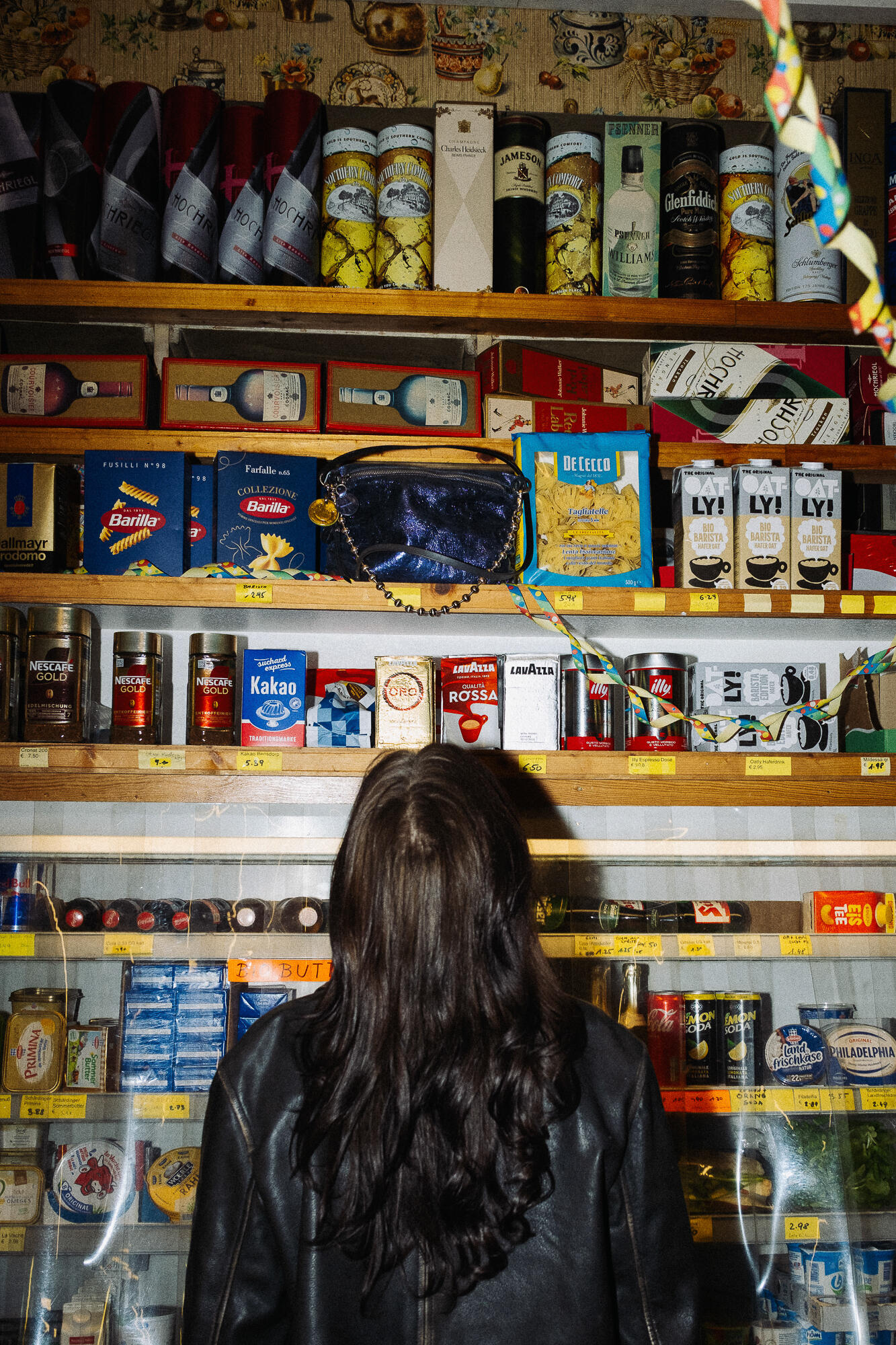 Eine Person mit langen dunklen Haaren und einer schwarzen INA KENT-Jacke steht in einem Geschäft vor Regalen, die mit verschiedenen Lebensmitteln und Getränken gefüllt sind.