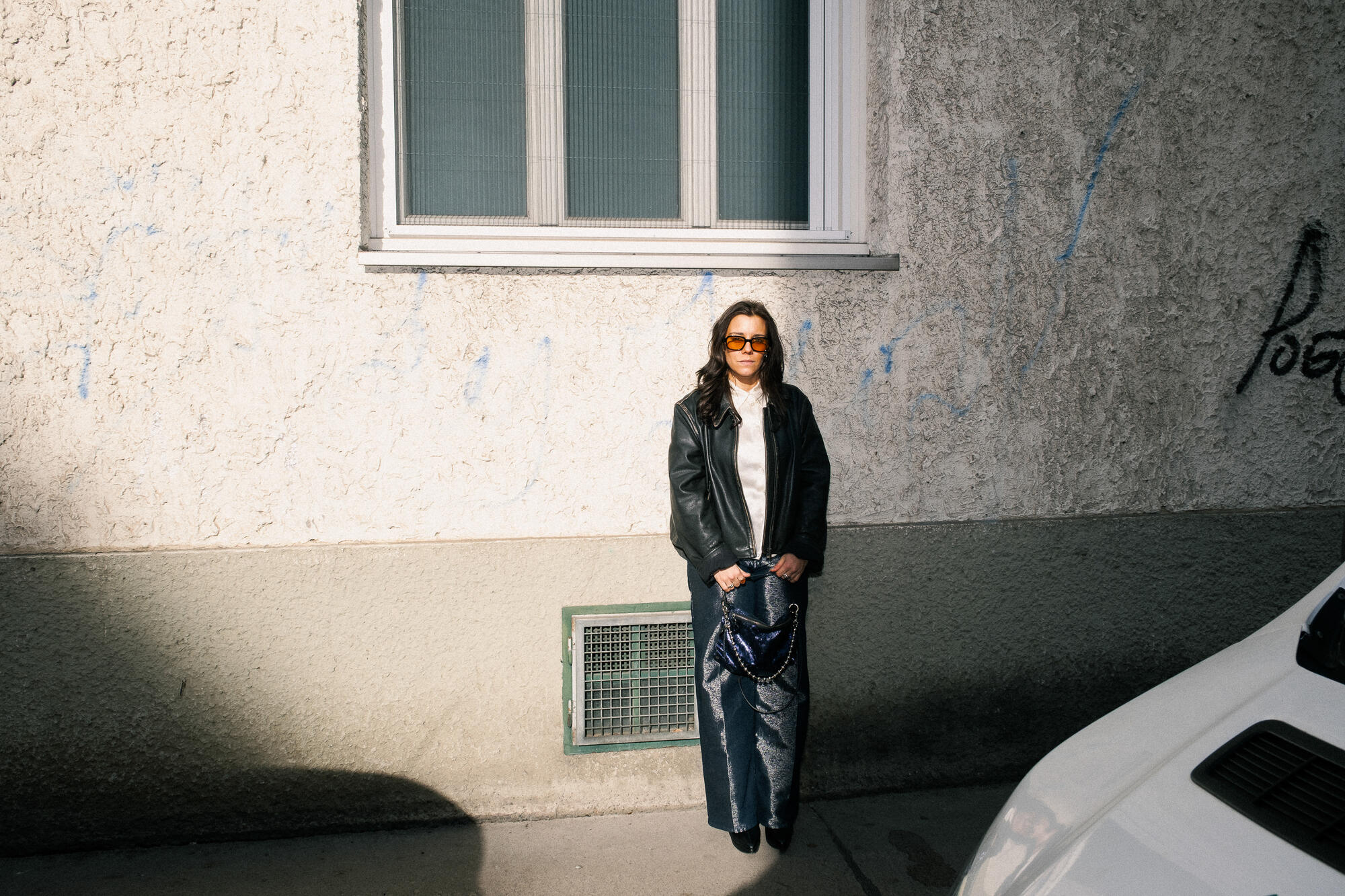 Eine Person mit Sonnenbrille, schwarzer Lederjacke und Jeans steht an einer strukturierten Wand unter einem Fenster und hält eine gemusterte INA KENT-Tasche.
