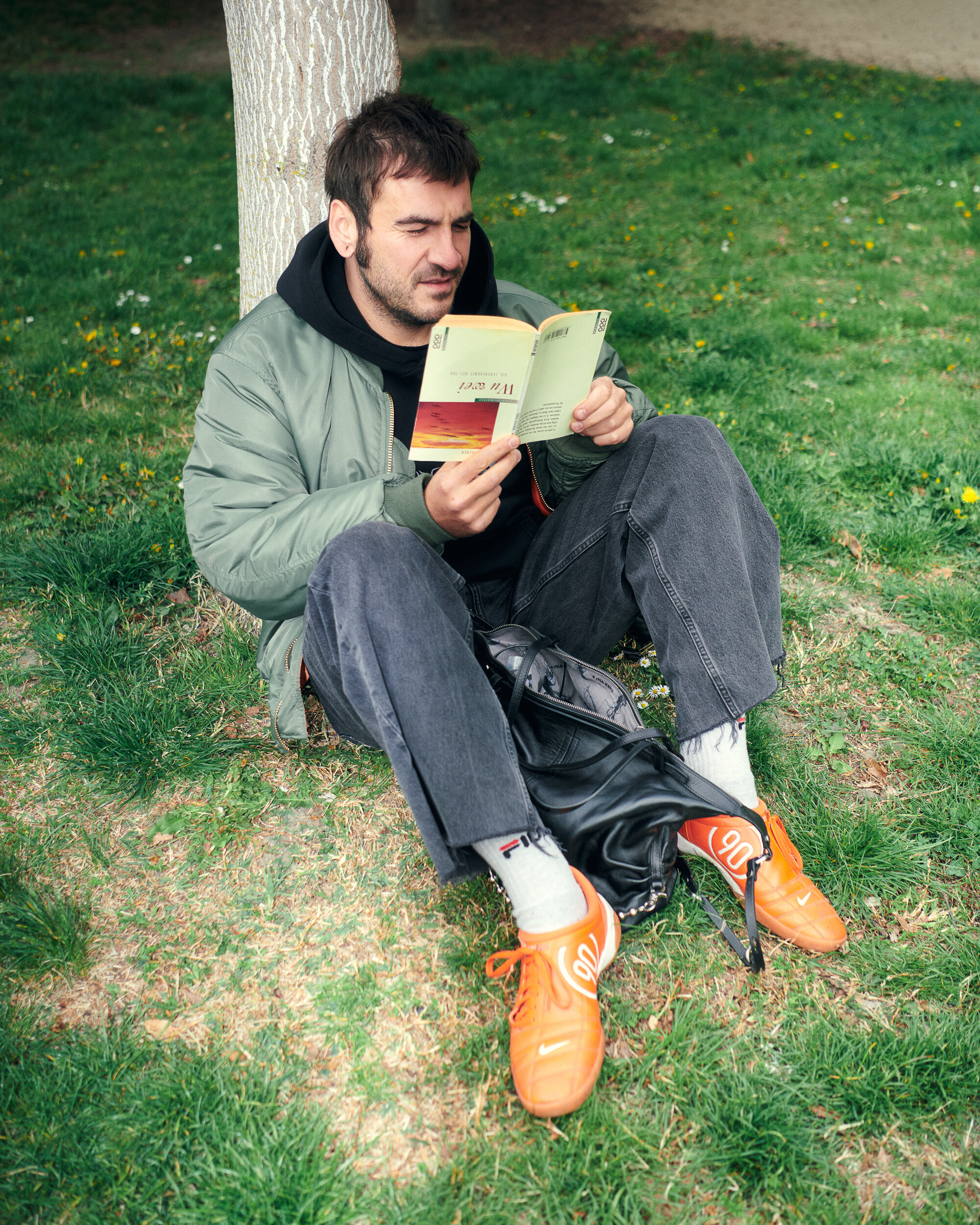 Ein Mann sitzt im Gras vor einem Baum und liest ein Buch. Er trägt eine olivfarbene Jacke, graue Jeans und orangefarbene Turnschuhe. Neben ihm auf dem Boden liegt eine schwarze INA KENT-Tasche.