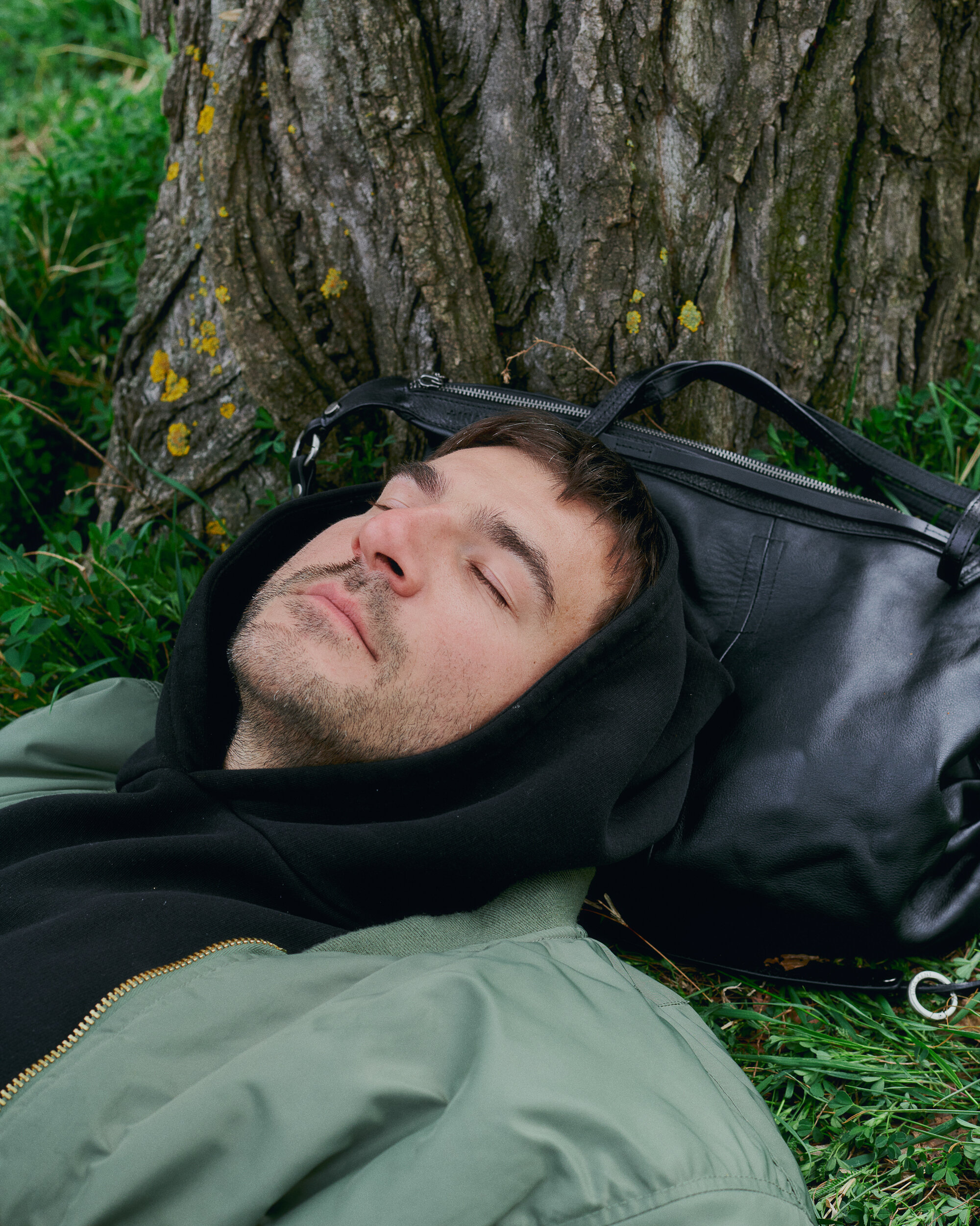 Eine Person in grüner Jacke und schwarzem Kapuzenpulli liegt mit geschlossenen Augen im Gras und stützt ihren Kopf auf eine schwarze INA KENT-Tasche an einem Baumstamm.