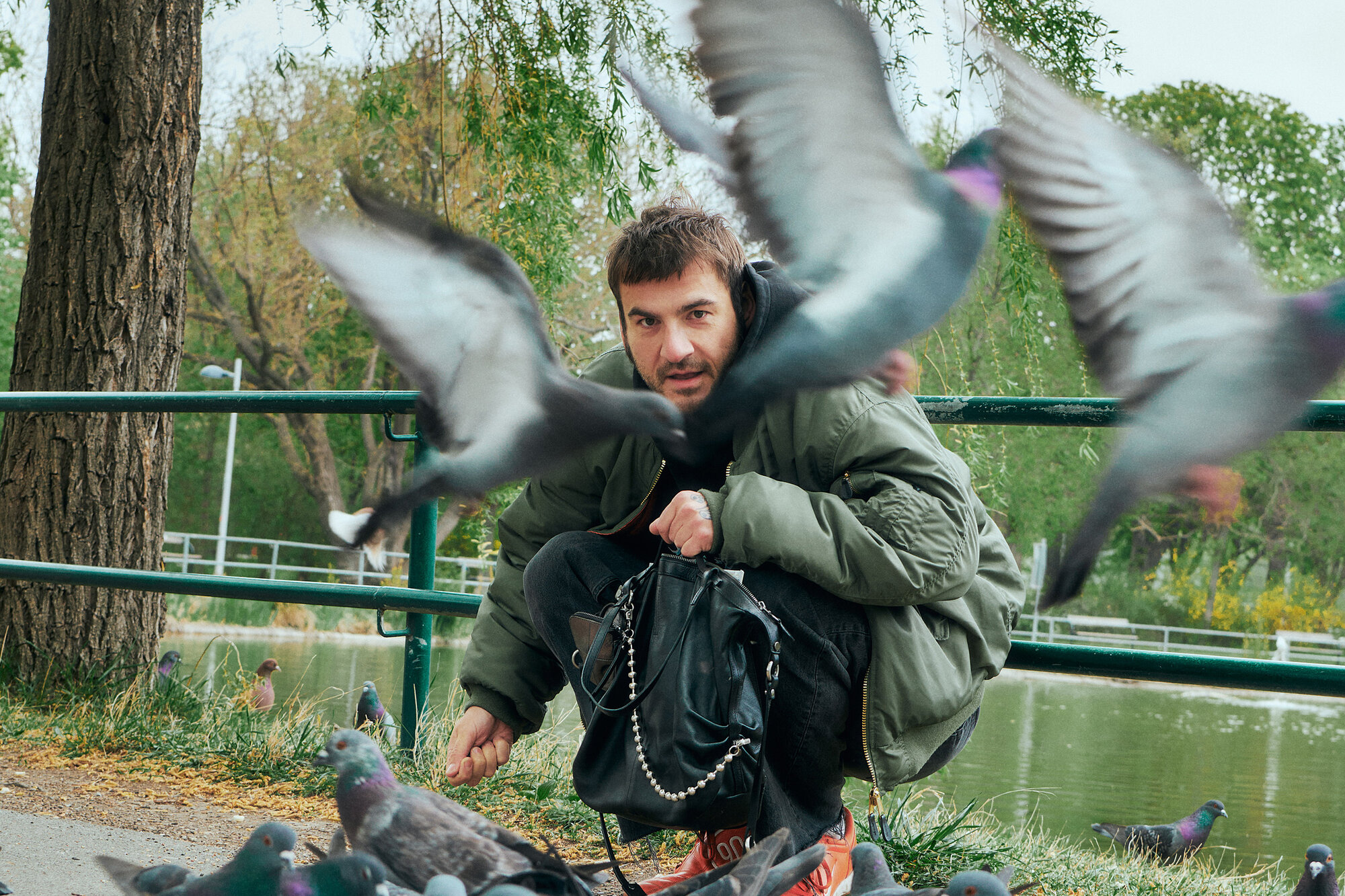 Ein Mann hockt in der Nähe eines Teiches, umgeben von Tauben, wobei mehrere Vögel im Flug sind - einer hält eine INA KENT-Tasche fest - und sein Gesicht teilweise verdecken.
