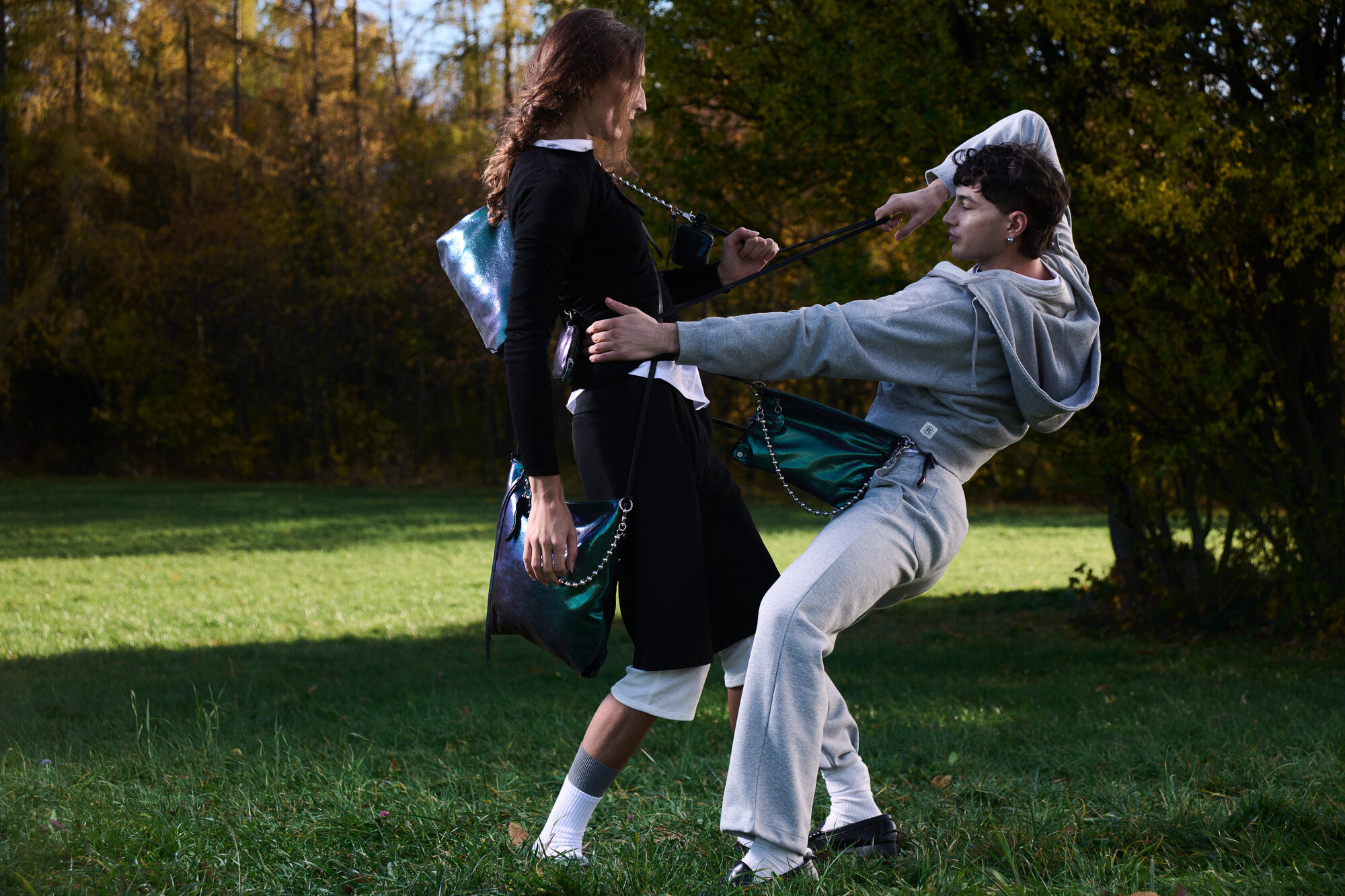 Two people outdoors on grass, playfully pulling each other by INA KENT bag straps—one leaning back, the other upright—amid trees with autumn foliage in the background.