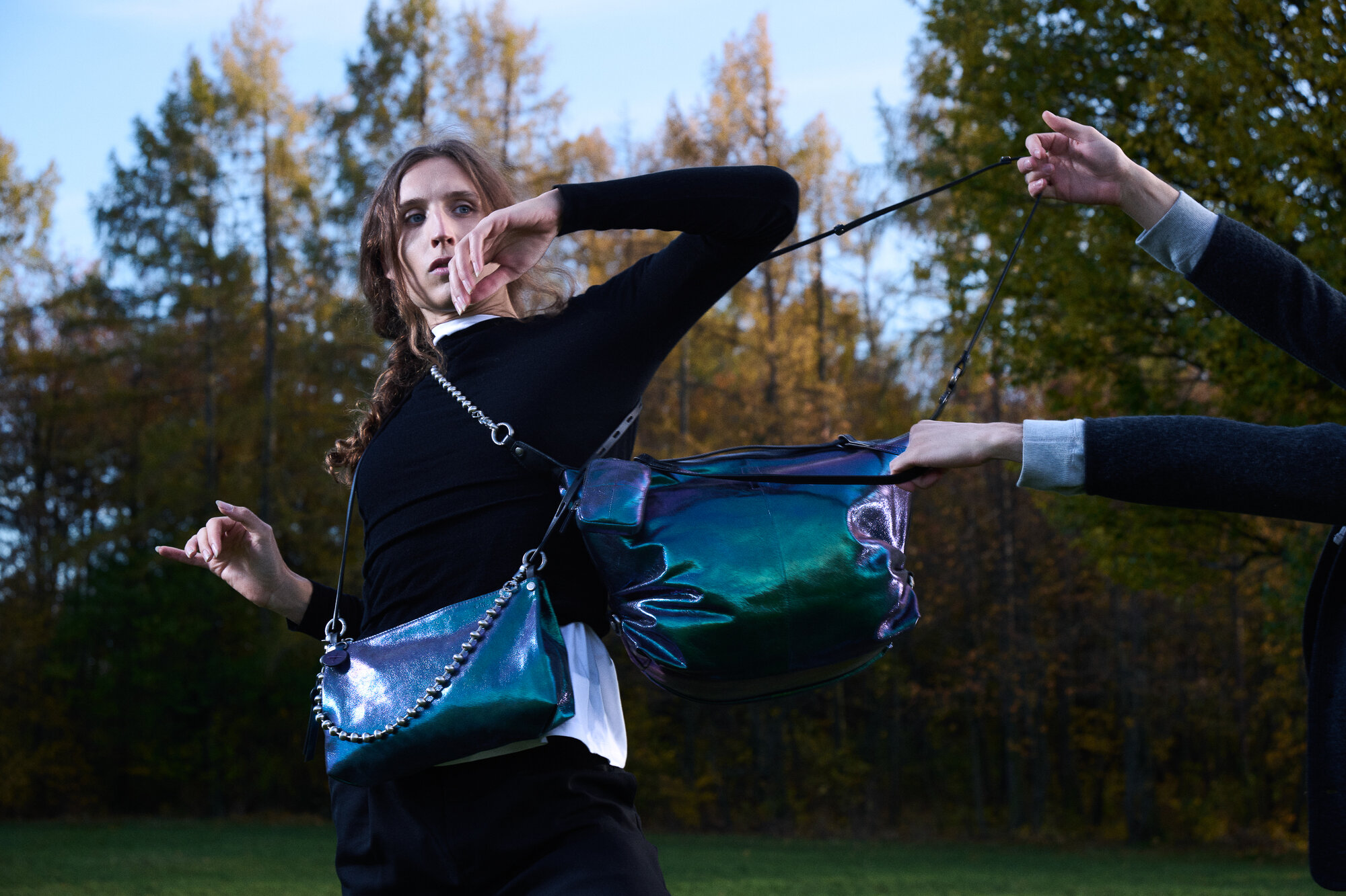 Eine schwarz-weiß gekleidete Person im Freien hält eine INA KENT-Metallic-Tasche, während eine andere Hand am Riemen der Tasche zieht. Im Hintergrund sind Bäume mit Herbstlaub zu sehen.