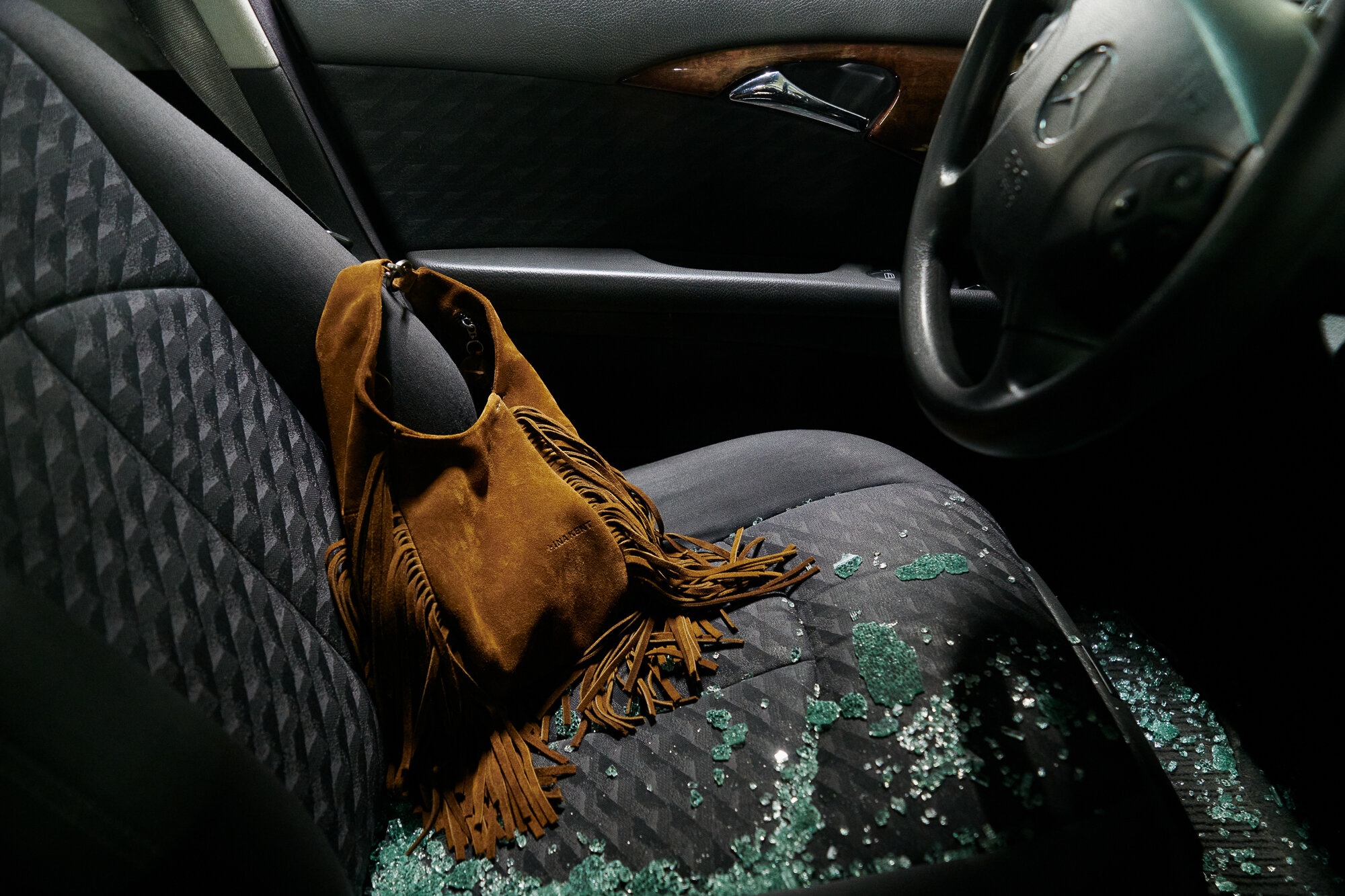 An INA KENT brown fringed purse sits on a car seat next to shattered glass, indicating a possible break-in through the passenger-side window.