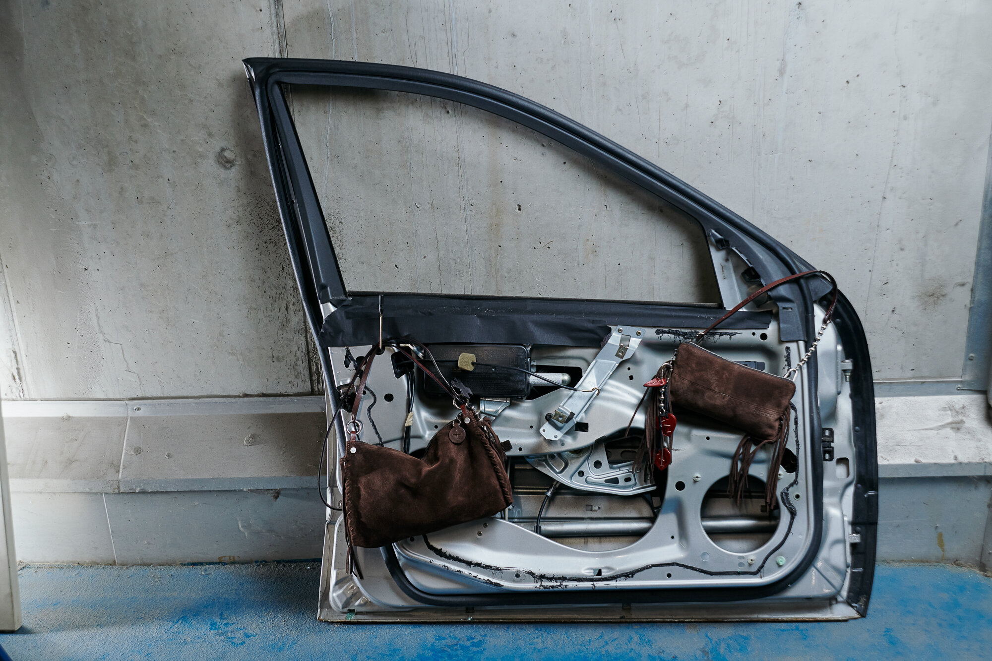 A detached car door leans against a concrete wall, with two brown INA KENT handbags hanging from the door’s inner metal frame.