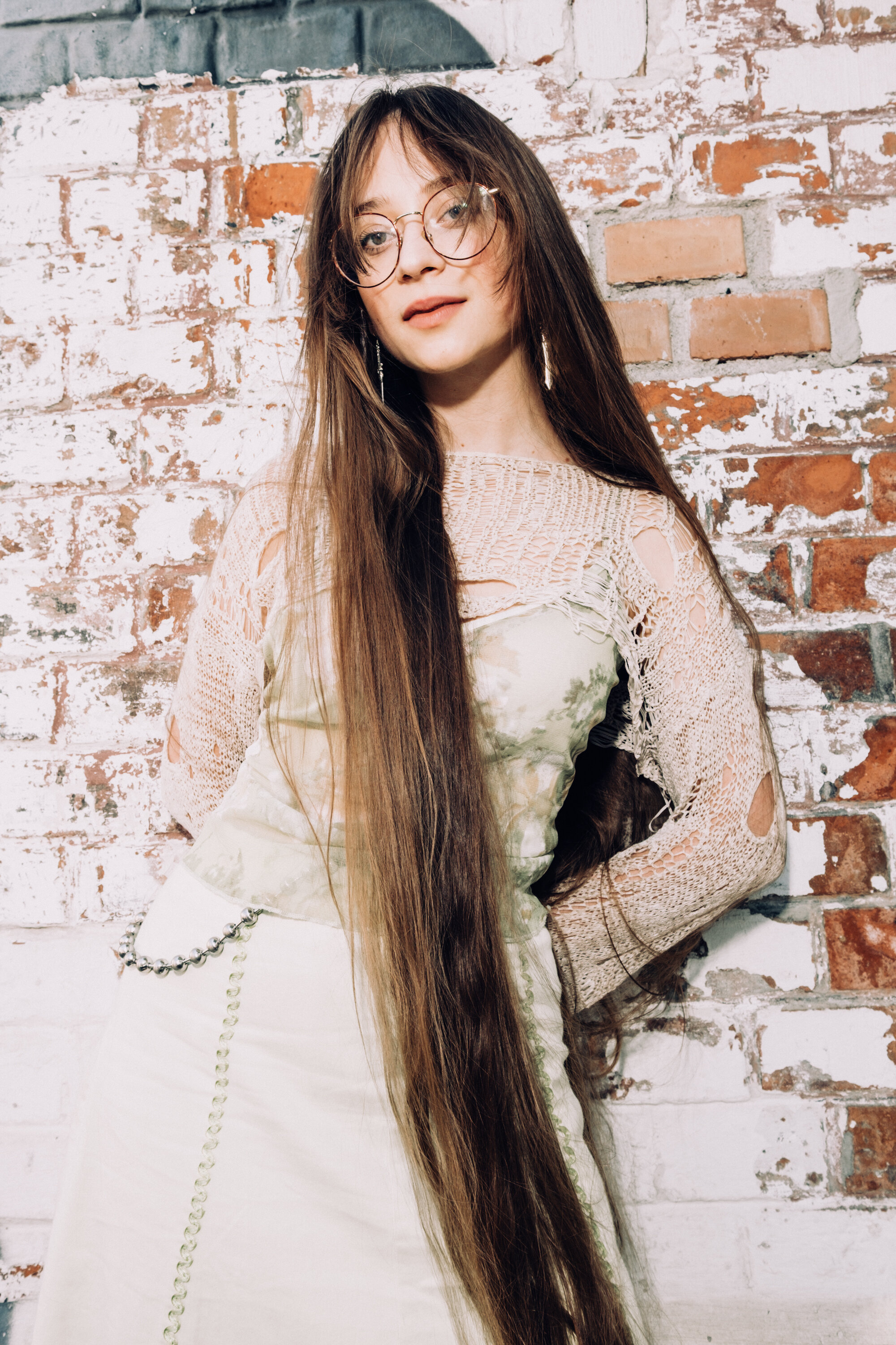 A young woman with very long brown hair, wearing glasses and a lace top, stands in front of a white brick wall with exposed red bricks, effortlessly stylish with her INA KENT bag slung over her shoulder.