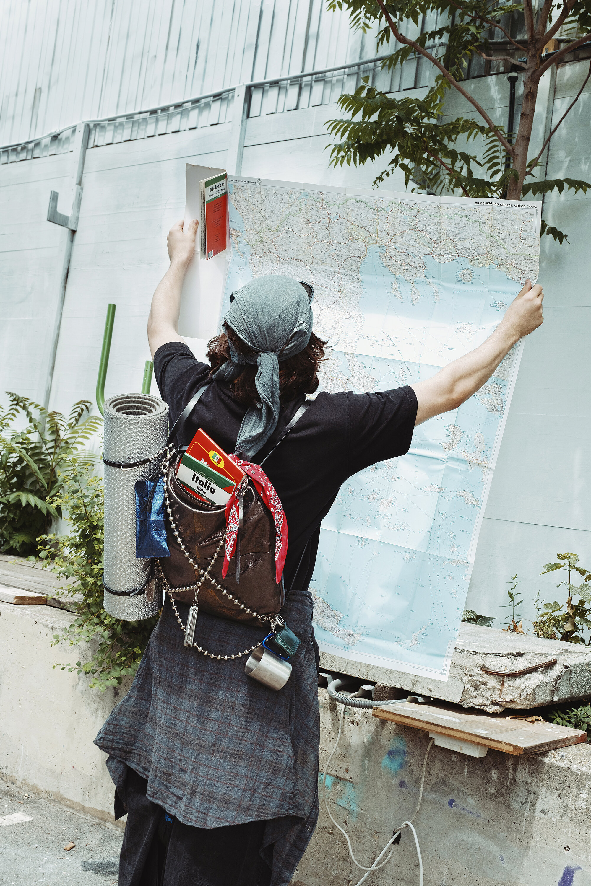 Person with an INA KENT backpack and headscarf stands outdoors holding a large map open, appearing to study it. Camping gear and a map labeled "Italia" are visible on their backpack.
