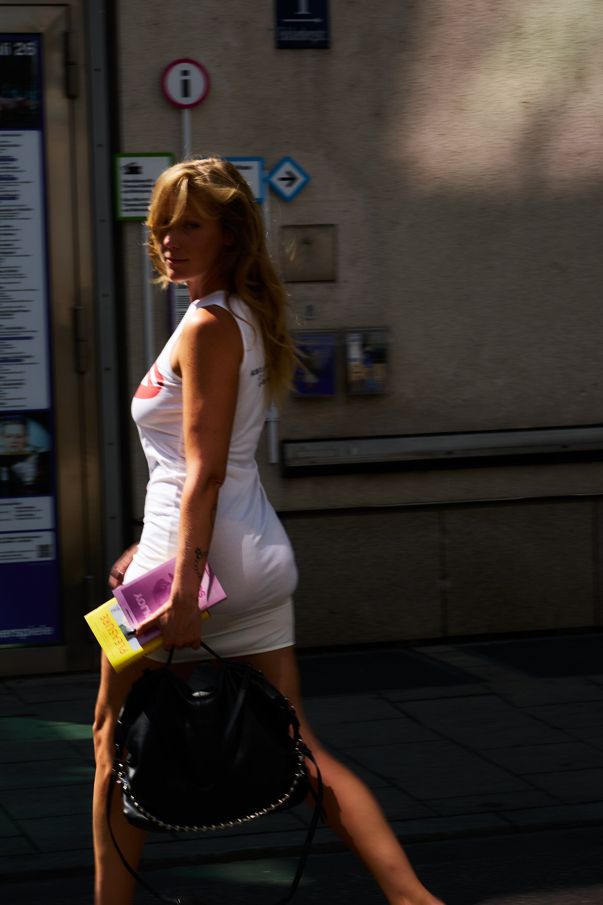 Eine Frau in einem weißen Kleid hält Bücher und eine schwarze INA KENT-Tasche in der Hand, während sie eine Straße in der Stadt entlanggeht und dabei über die Schulter zurückblickt.