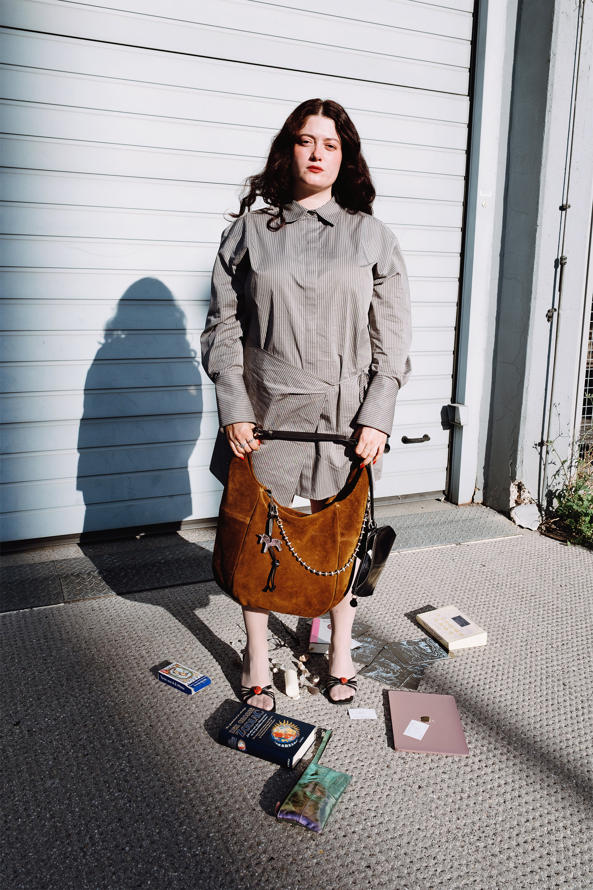 Eine Frau in grauer Kleidung steht mit einer großen, hellbraunen INA KENT-Tasche auf dem Bürgersteig; um sie herum liegen Bücher und ein Laptop auf dem Boden verstreut, vor einem geschlossenen Industrietor.
