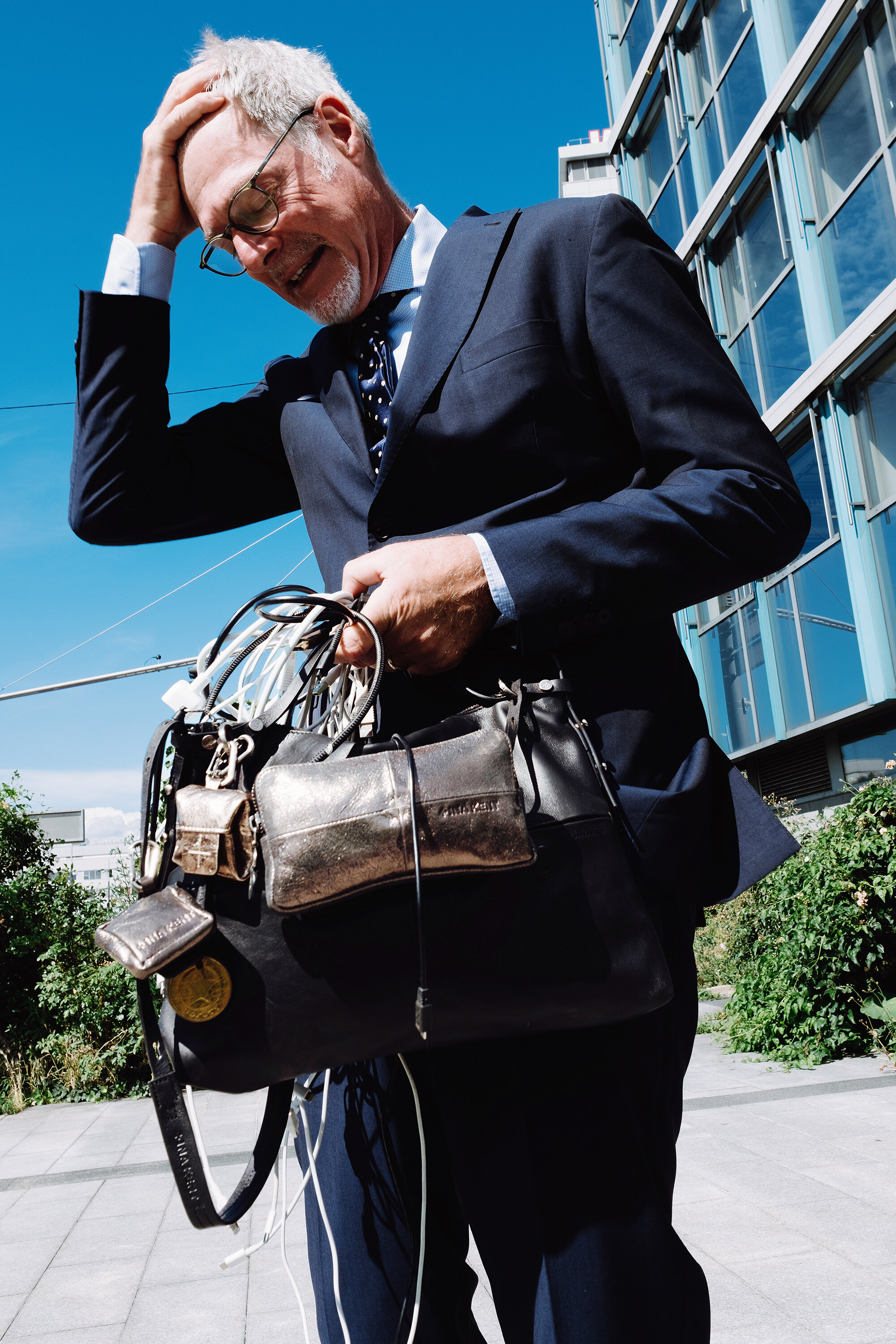 Ein Mann im Anzug steht im Freien, hält eine große INA KENT-Handtasche voller verschiedener Gegenstände und Kabel in der Hand, während er sich an den Kopf fasst und nach unten schaut.