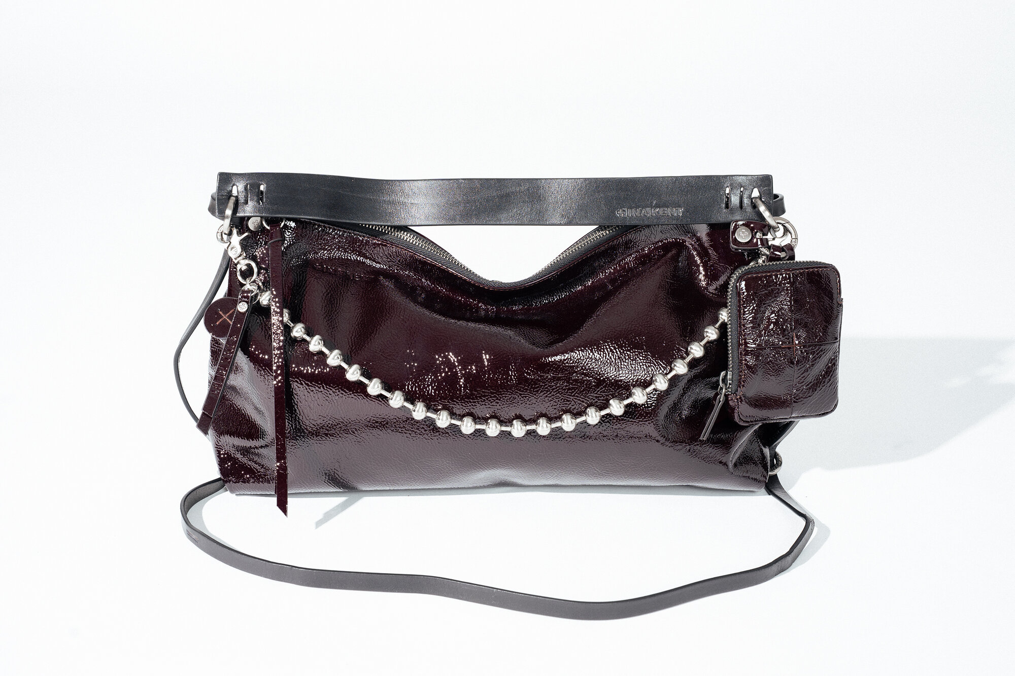 Eine glänzende, dunkelburgunderrote INA KENT-Handtasche mit einem schwarzen Riemen, einem metallischen Kettenelement und einem kleinen, farblich passenden Beutel, der daran befestigt ist.