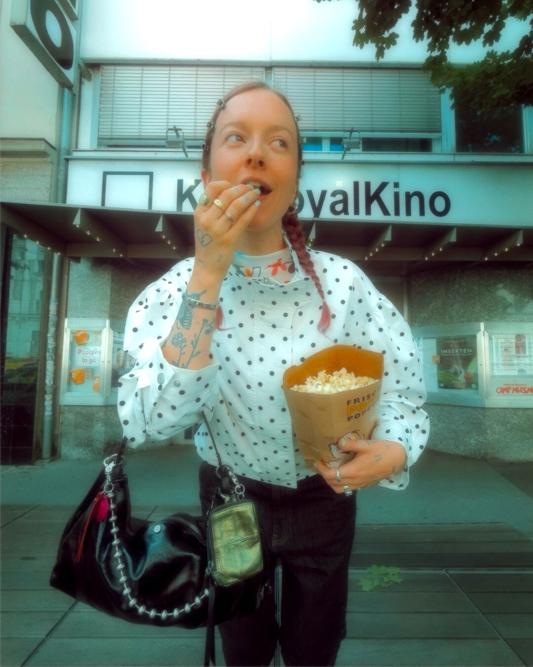 Eine Person mit Zöpfen isst vor einem Kino Popcorn; sie trägt ein gepunktetes Hemd und hält eine schwarze Handtasche von INA KENT in der Hand.