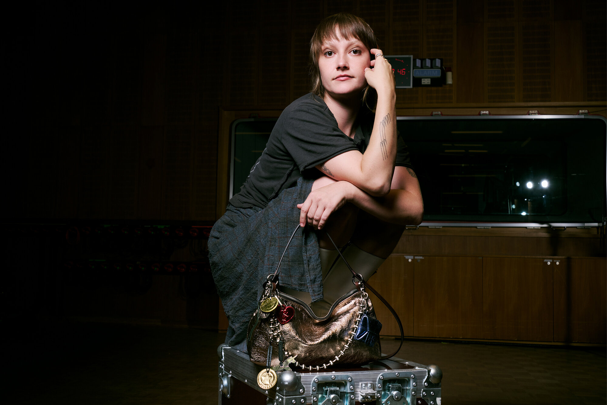 A person in casual clothing poses with one arm on their knee and the other holding an INA KENT metallic handbag, sitting on a large equipment case in a dimly lit room.