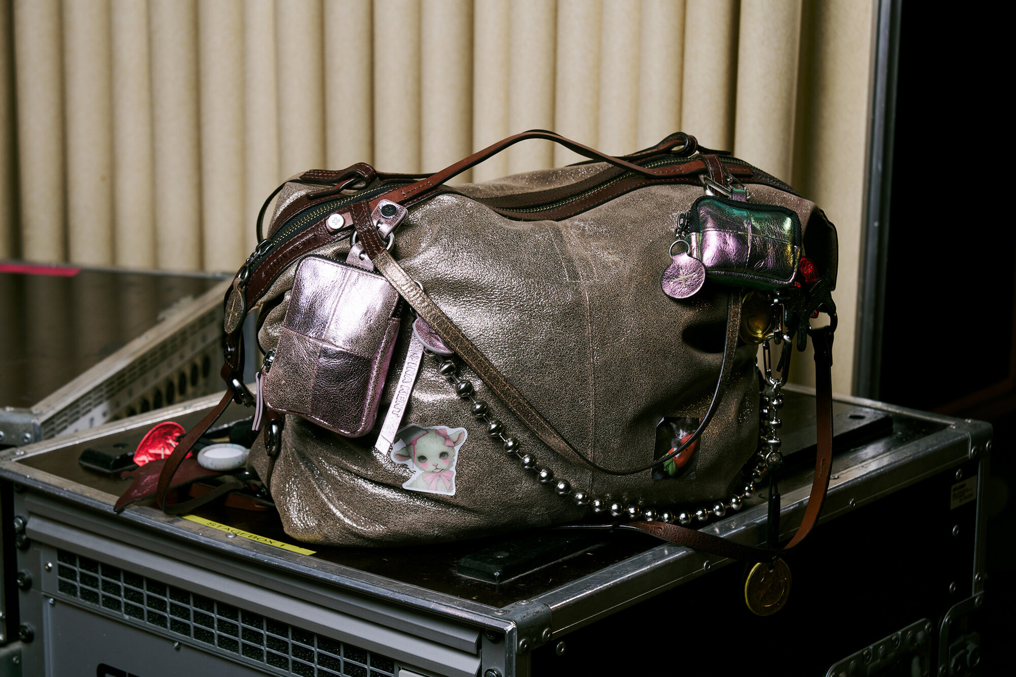 A large brown leather bag by INA KENT, adorned with multiple pockets, keychains, a cat sticker, and silver beads, sits on top of metal cases in front of beige vertical blinds.