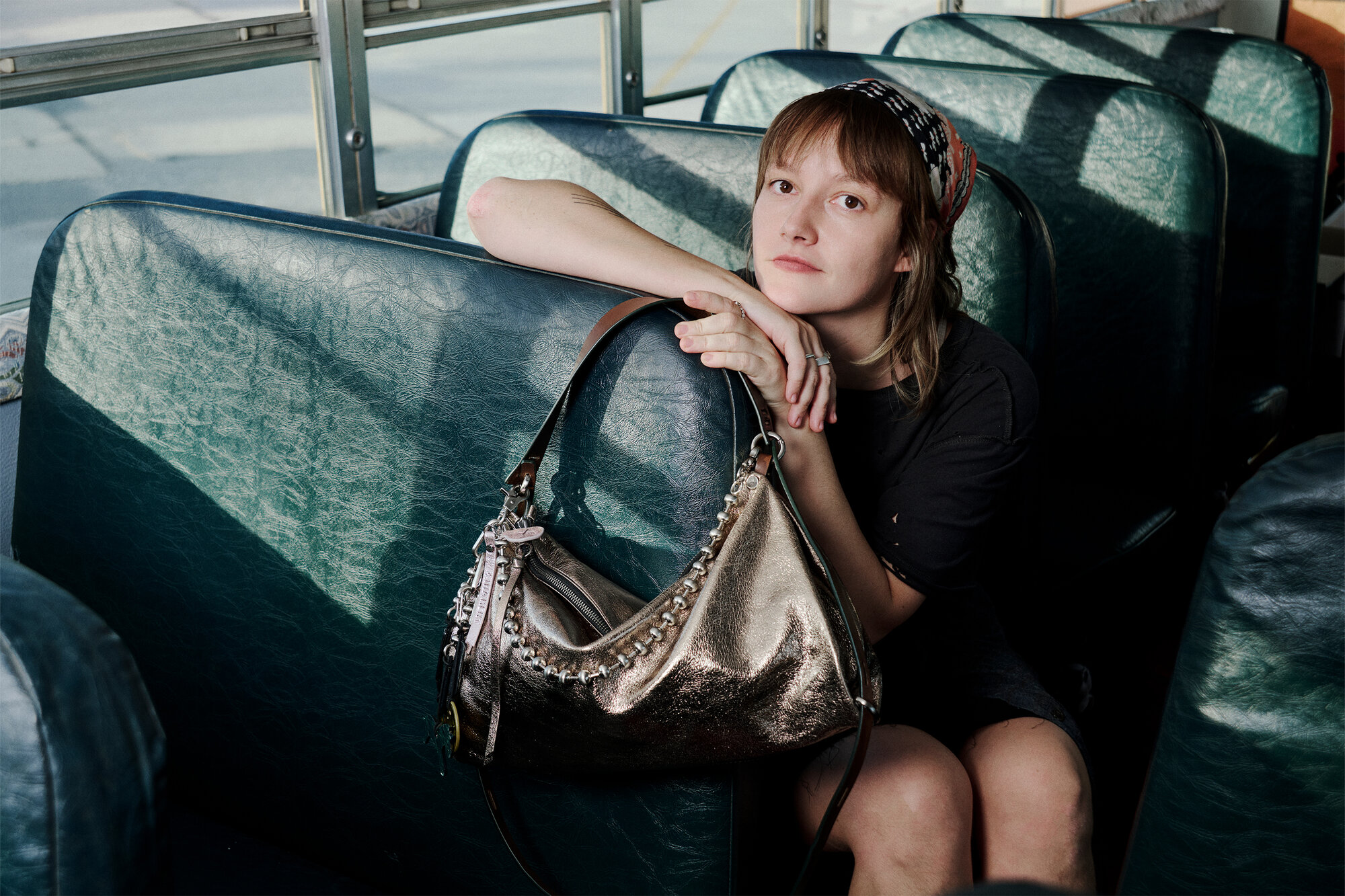 Ein junger Mensch sitzt allein auf einem Bussitz, die Arme auf der Rückenlehne abgestützt, neben sich auf dem Sitz liegt eine elegante metallische Handtasche von INA KENT.
