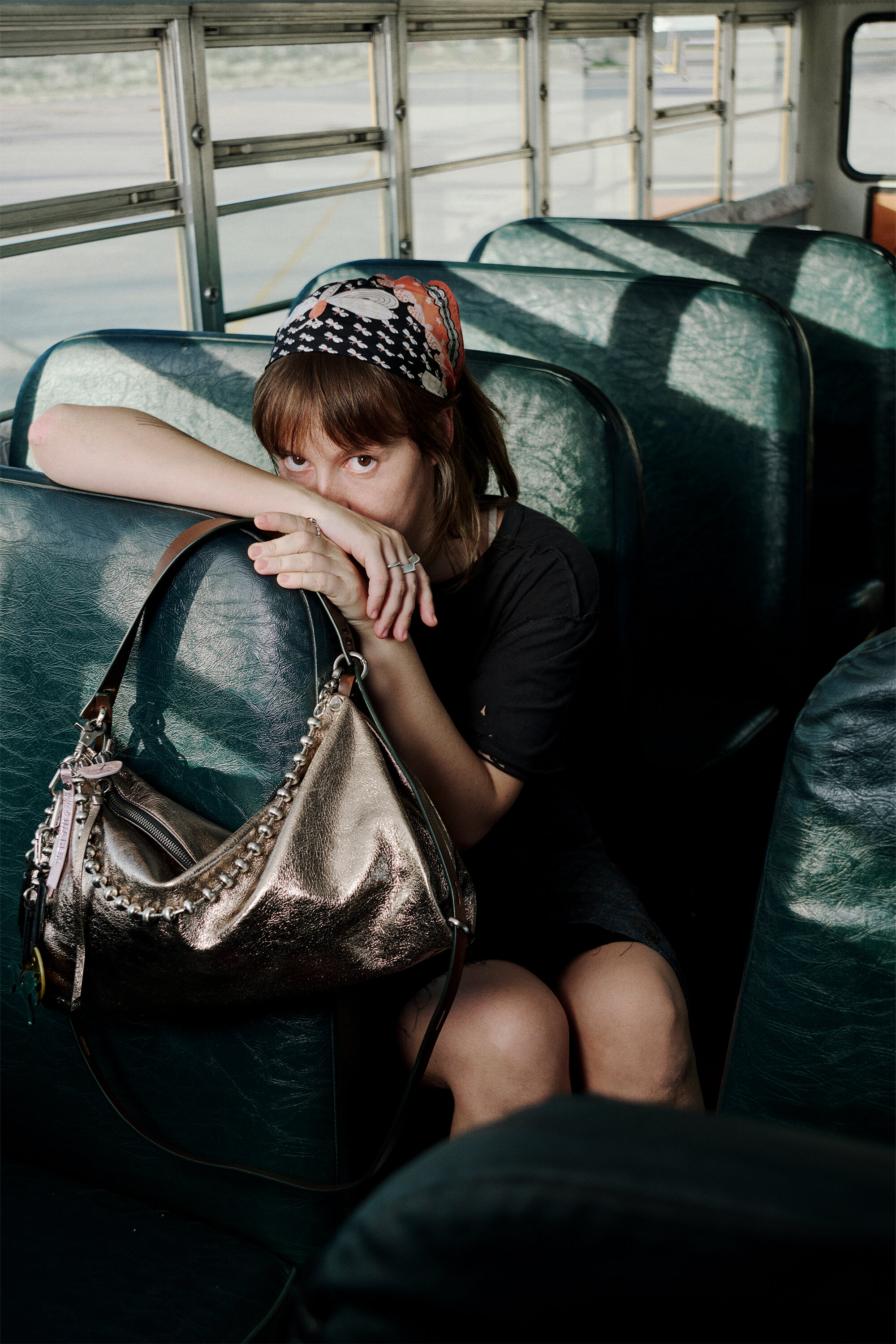 Eine Frau mit Kopftuch sitzt allein auf einem grünen Bussitz, den Arm auf dem Vordersitz abgestützt, während sie eine elegante, metallisch schimmernde INA KENT-Handtasche in der Hand hält.