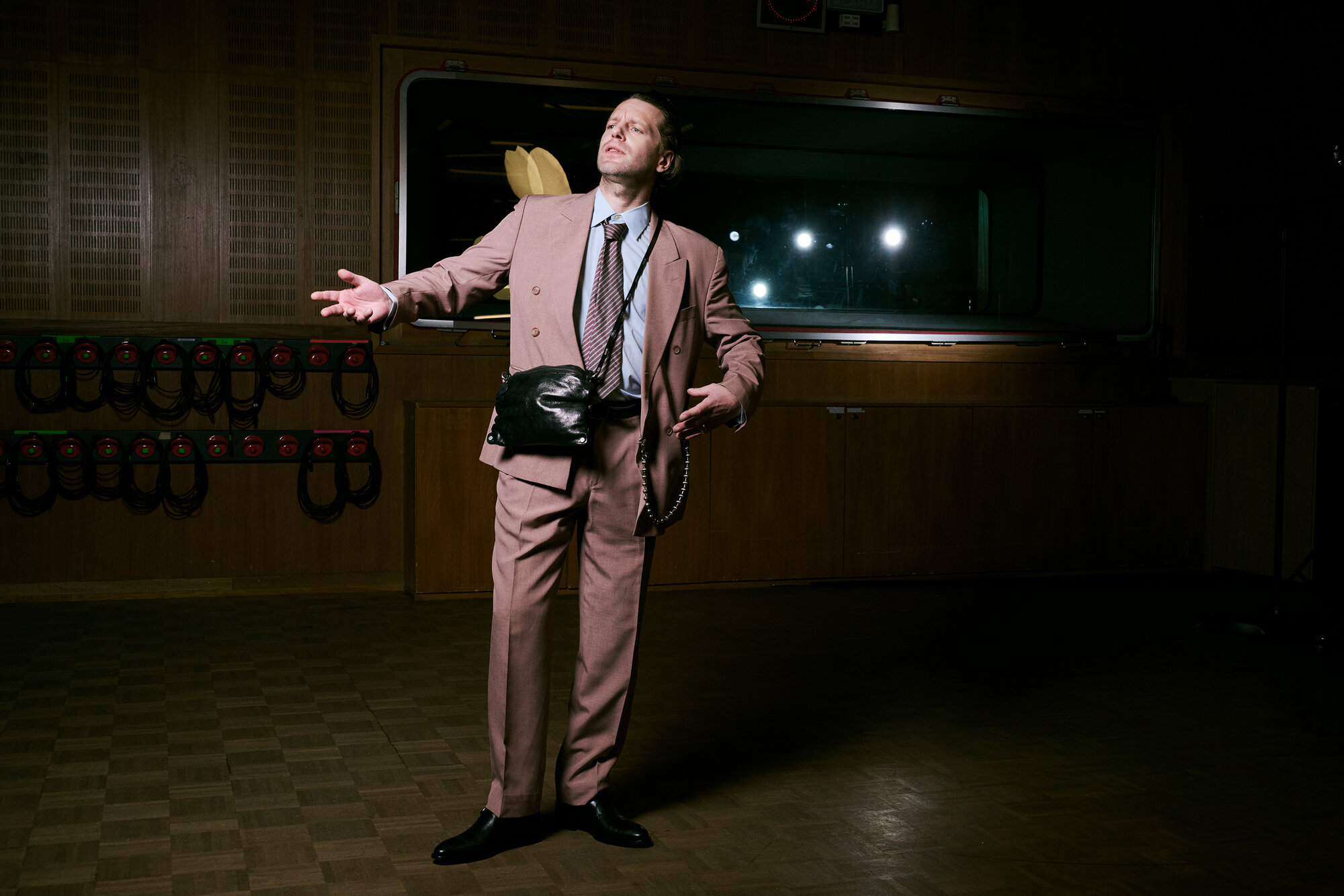 A man in a pink suit stands in a dimly lit room with wooden walls, one hand outstretched and an INA KENT black bag slung across his body.