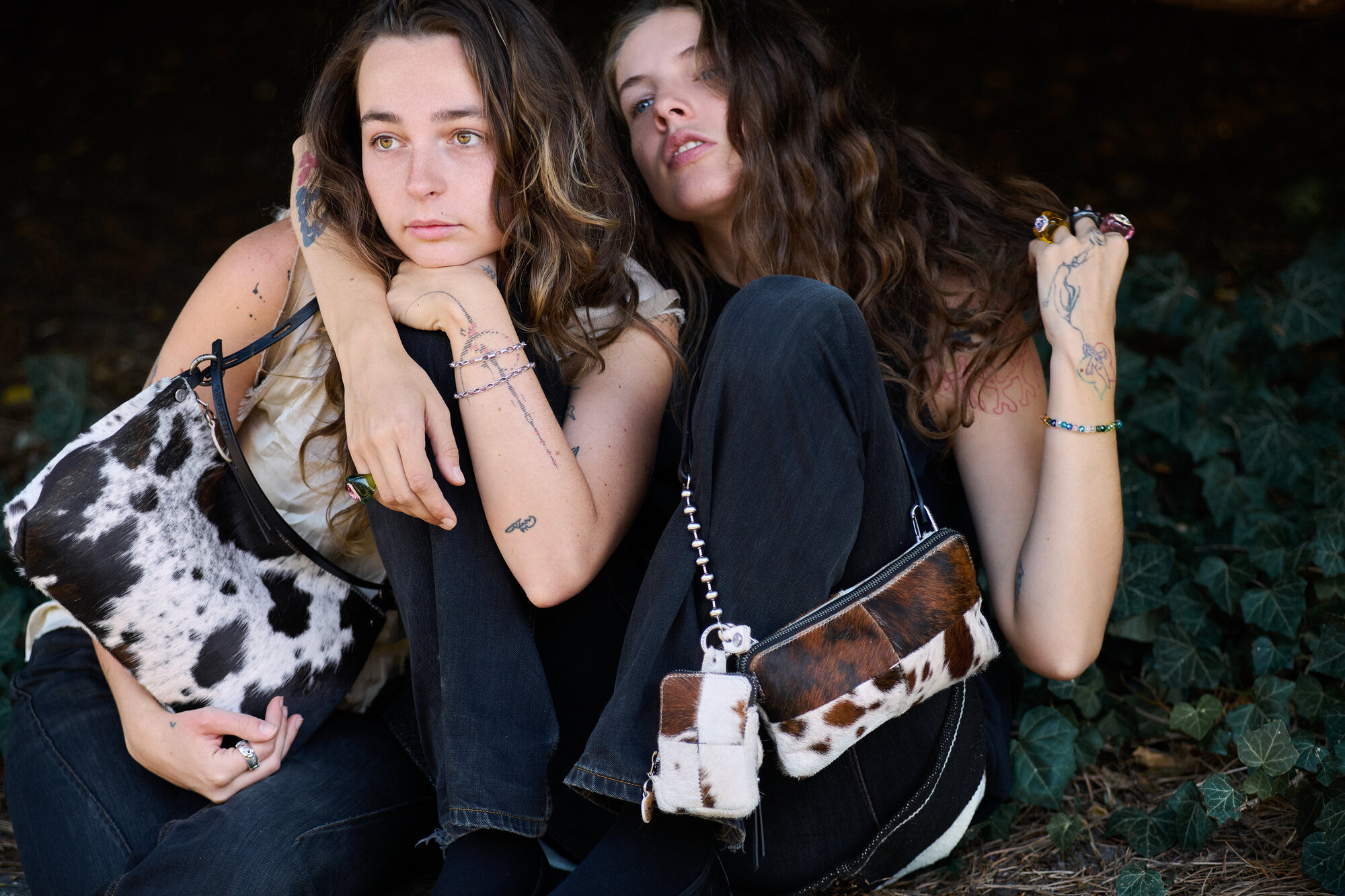 Two women with long hair sit closely outdoors, each holding an INA KENT cow-print handbag. Both sport casual clothes, visible tattoos, and rings.