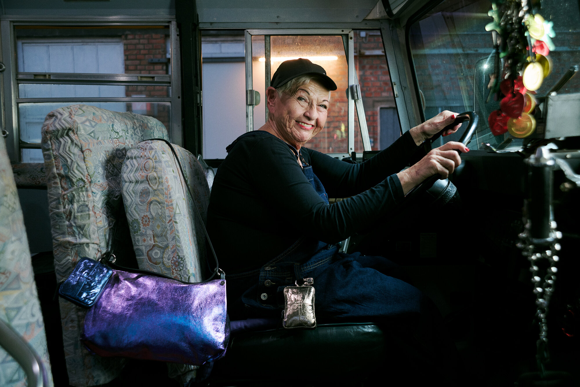 An older woman wearing a cap and dark clothing sits in the driver’s seat of a bus, smiling at the camera. Beside her, two shiny INA KENT bags are hanging, catching the light.