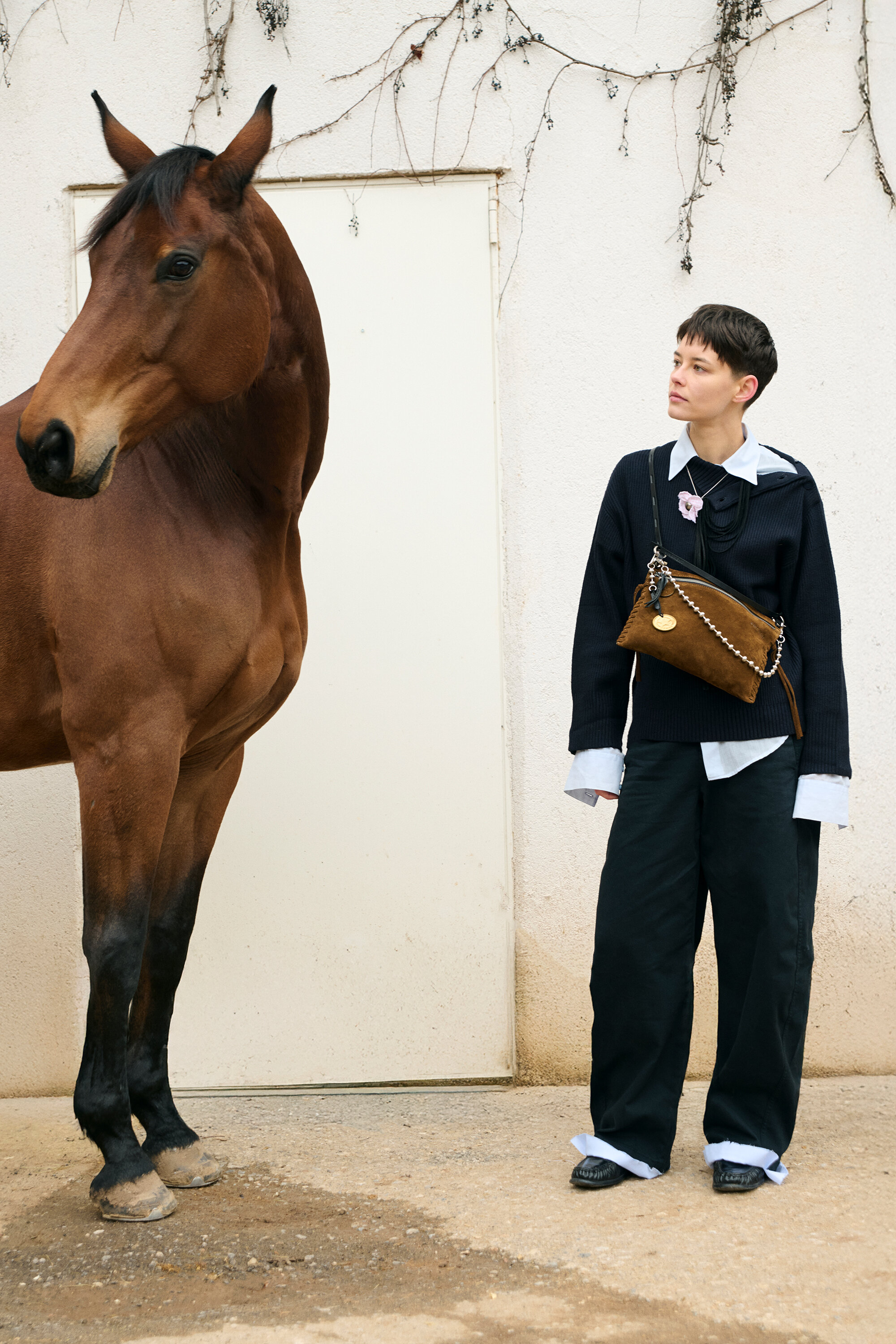 A person wearing loose black pants and an INA KENT sweater stands next to a brown horse in front of a white wall with some vines.