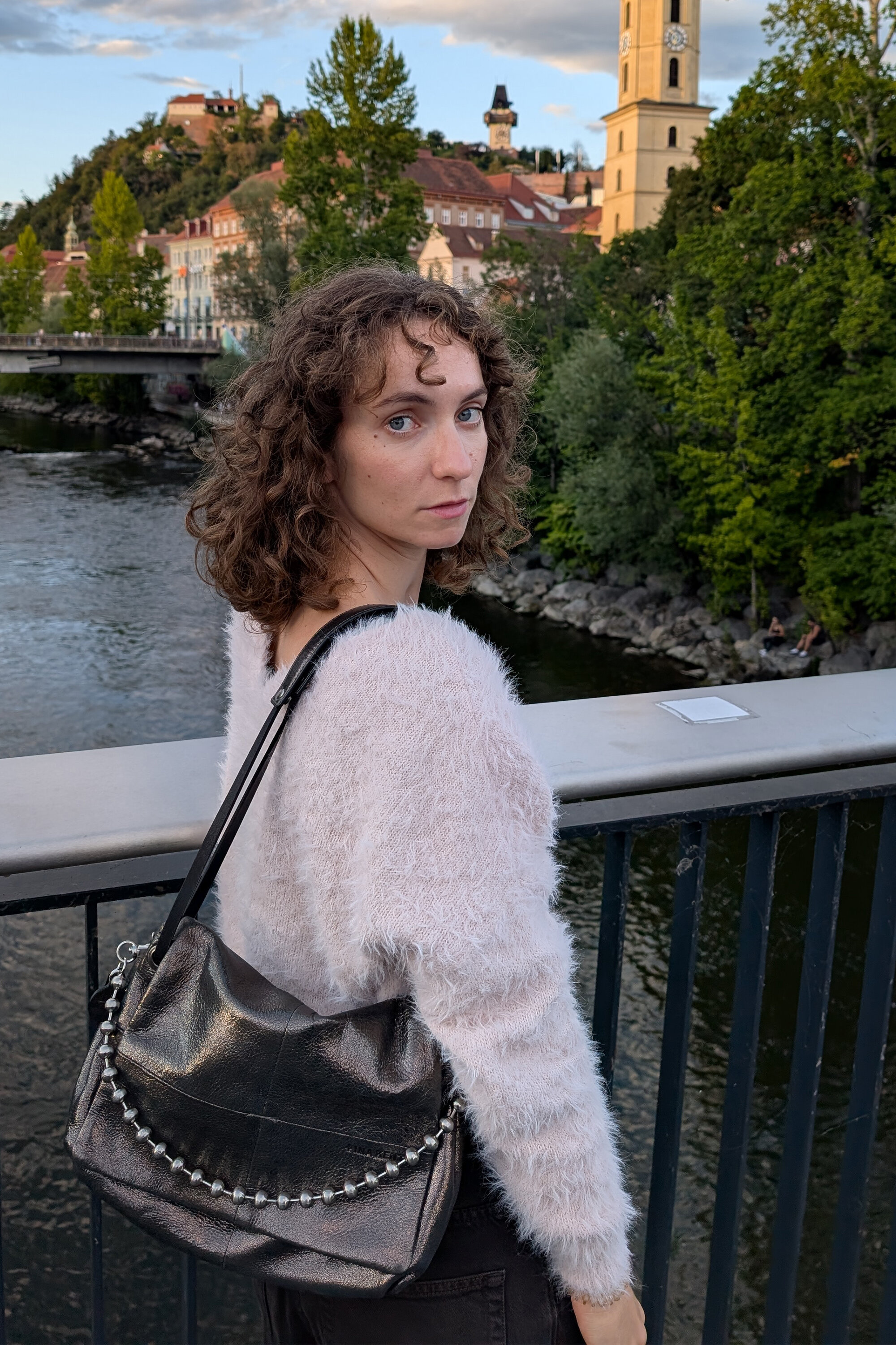 Eine Frau mit lockigem Haar, die eine INA KENT-Tasche trägt, steht auf einer Brücke mit Blick auf einen Fluss und eine Stadtlandschaft.