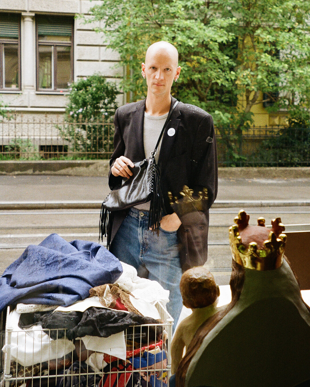 Eine Person mit rasiertem Kopf, die einen Blazer von INA KENT und Jeans trägt, steht auf einem Bürgersteig in der Stadt neben einem Korb mit Stoff und einer Statue einer Figur mit Krone.